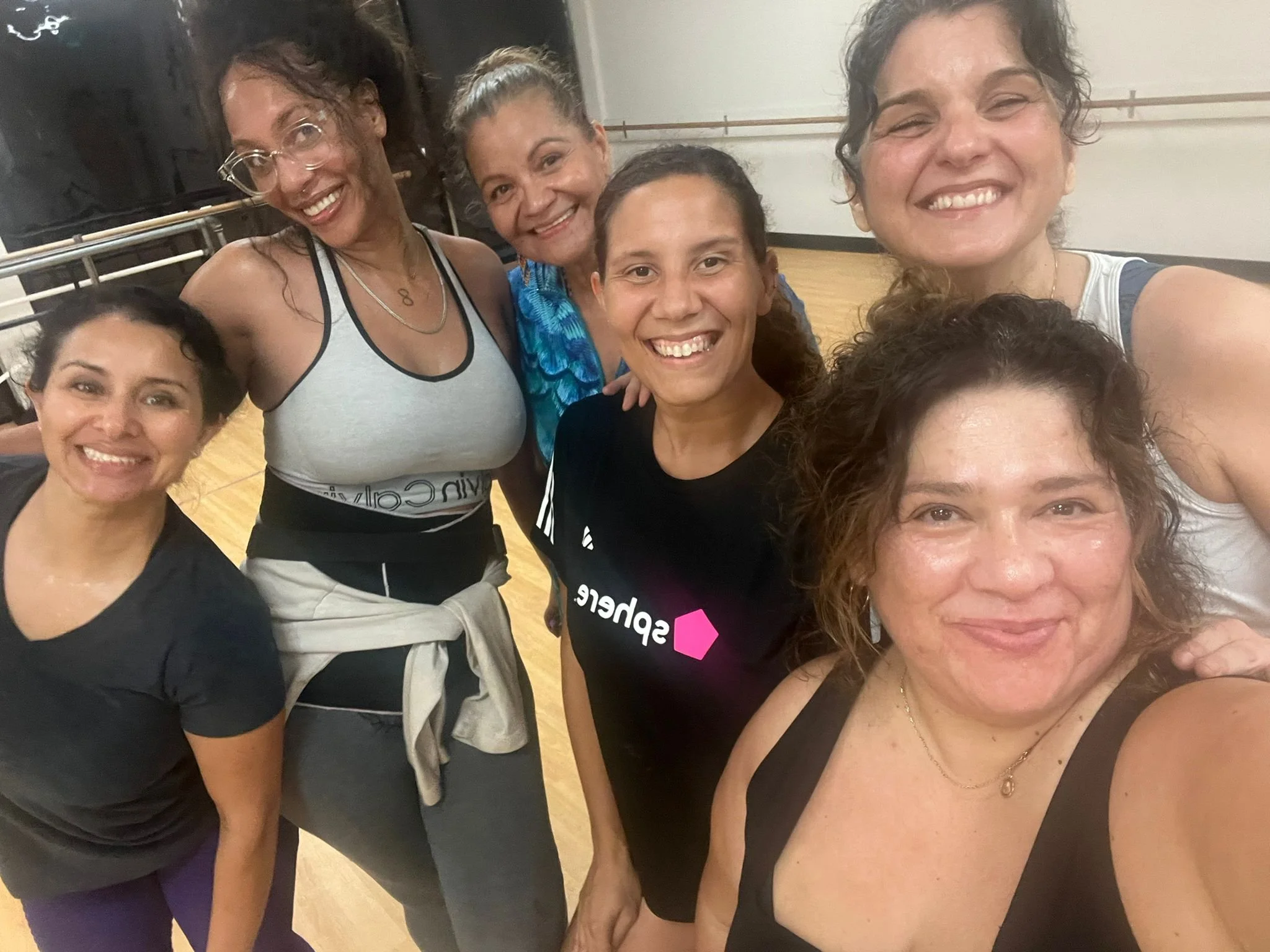 Group of seven women smiling and taking a selfie in a dance or fitness studio.