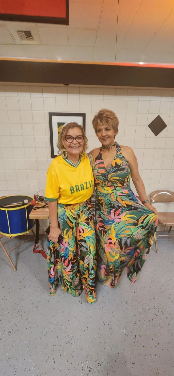 Two women with short curly hair, smiling, wearing colorful floral skirts, and one wearing a yellow Brazil soccer jersey. They are standing indoors against a white tiled wall with framed art, and there are chairs and a drum in the background.