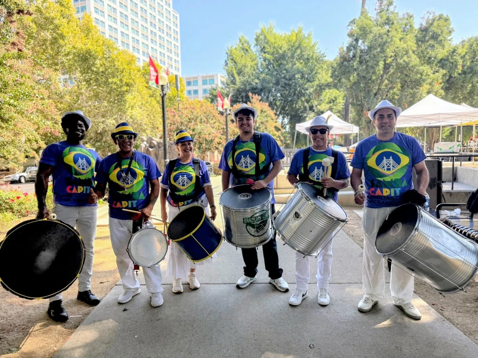 Six people dressed in blue Brazil-themed shirts with drums and percussion instruments, standing outdoors on a bright day, with trees, flags, and buildings in the background.  G.R.E.S. unidos da capital of sacramento brazilian samba performing arts
