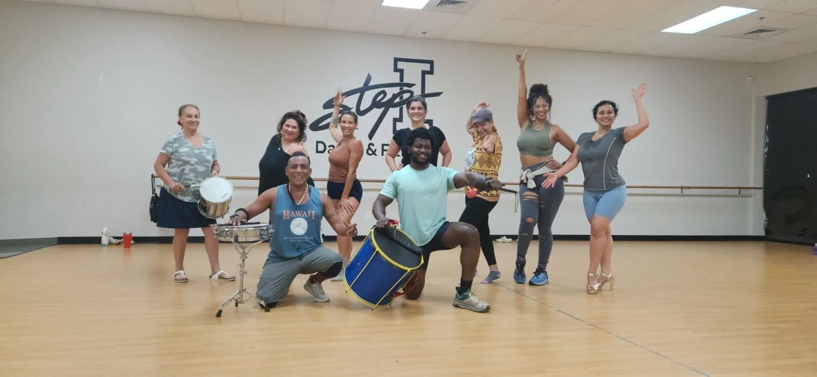 Group of eight diverse people in a dance studio posing with musical instruments, all smiling and some raising their hands, with the studio's logo visible on the wall behind them. Brazilian samba class sacramento dance