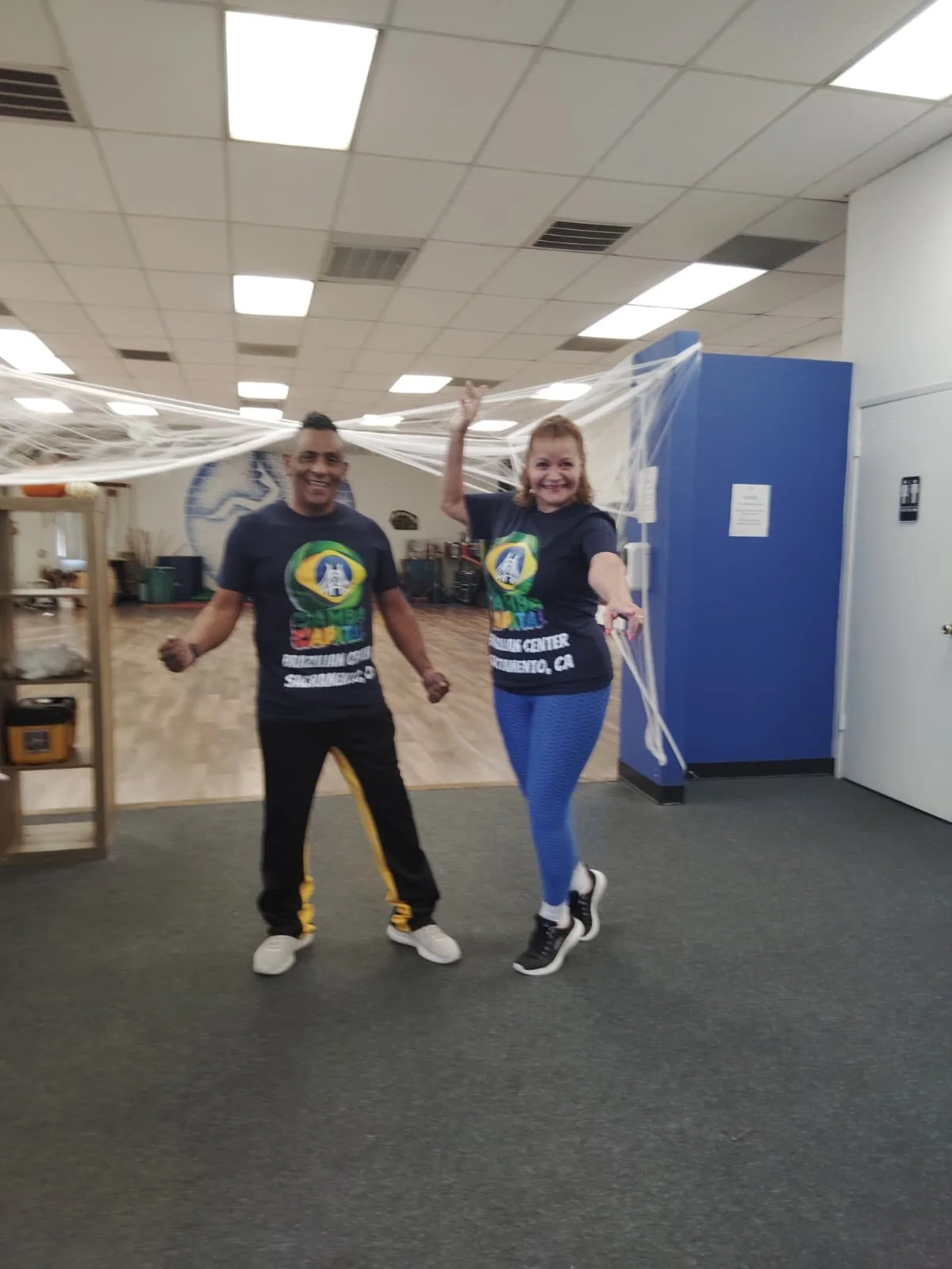 Two smiling people wearing matching t-shirts, standing inside a room decorated with fake spider webs, celebrating or participating in an event.