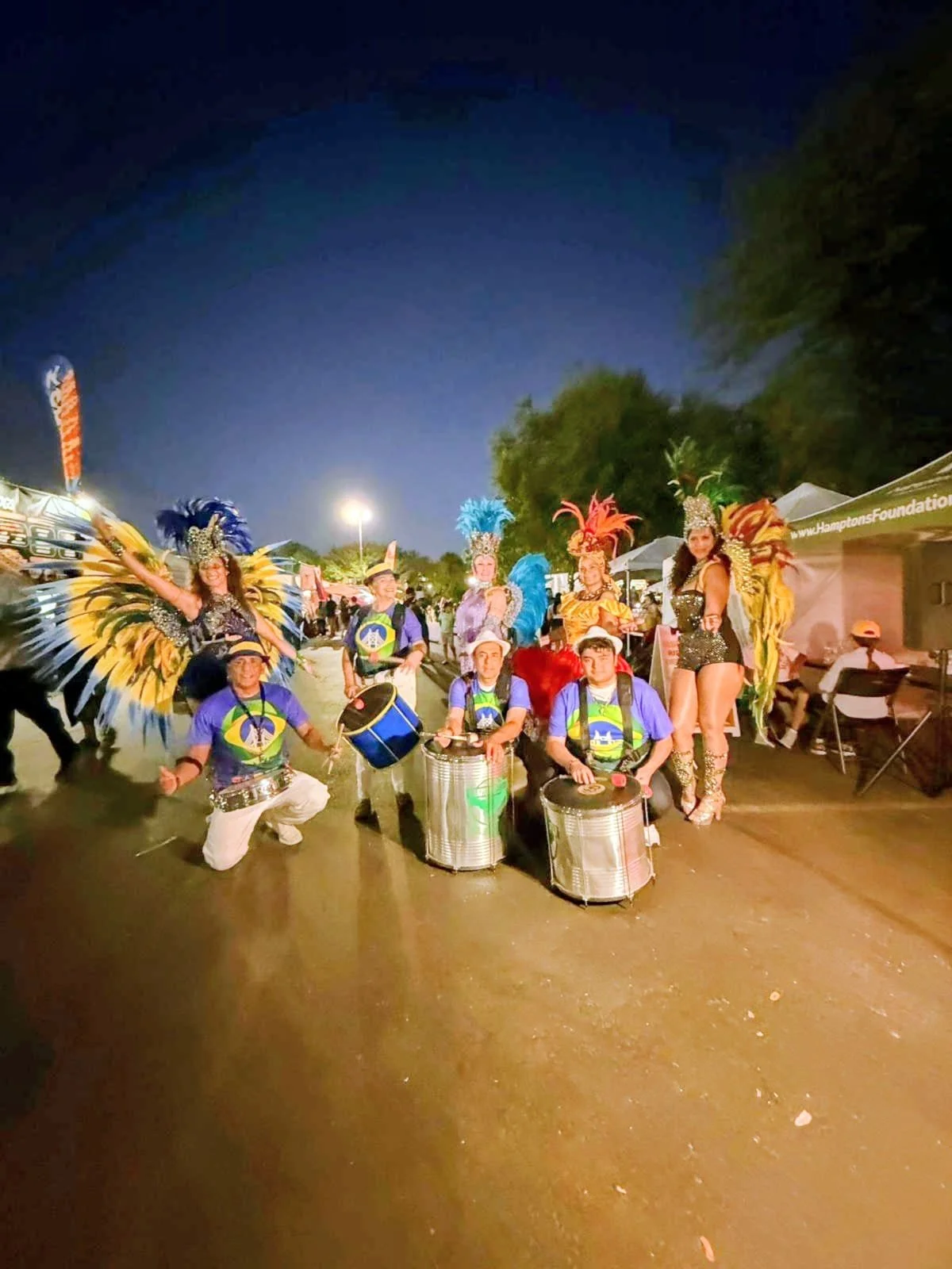 Group of performers dressed in colorful carnival costumes with feathered headdresses, standing with musical instruments at night during a festival.