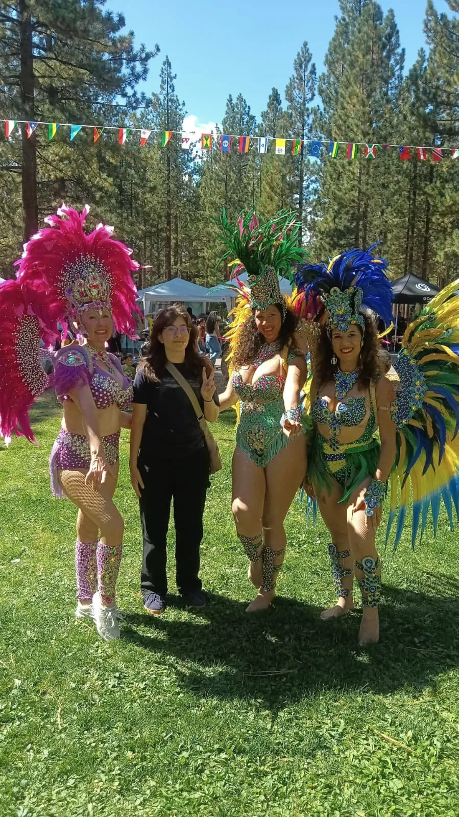Four women dressed in colorful carnival costumes with feathers and beads, standing outdoors on grass with trees and a blue sky in the background.