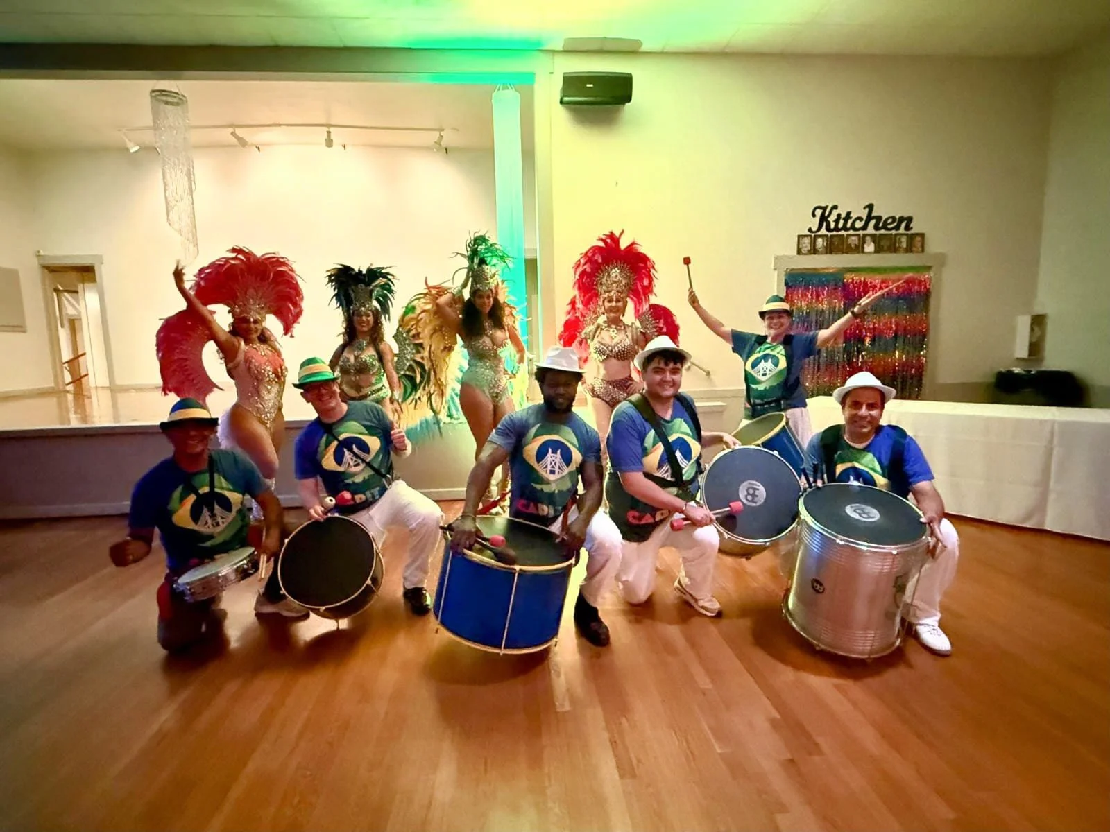 A group of five musicians playing drums in front of four showgirls with feathered headdresses, colorful costumes, and dancing poses, in a decorated indoor venue with a sign that says "Kitchen" in the background.