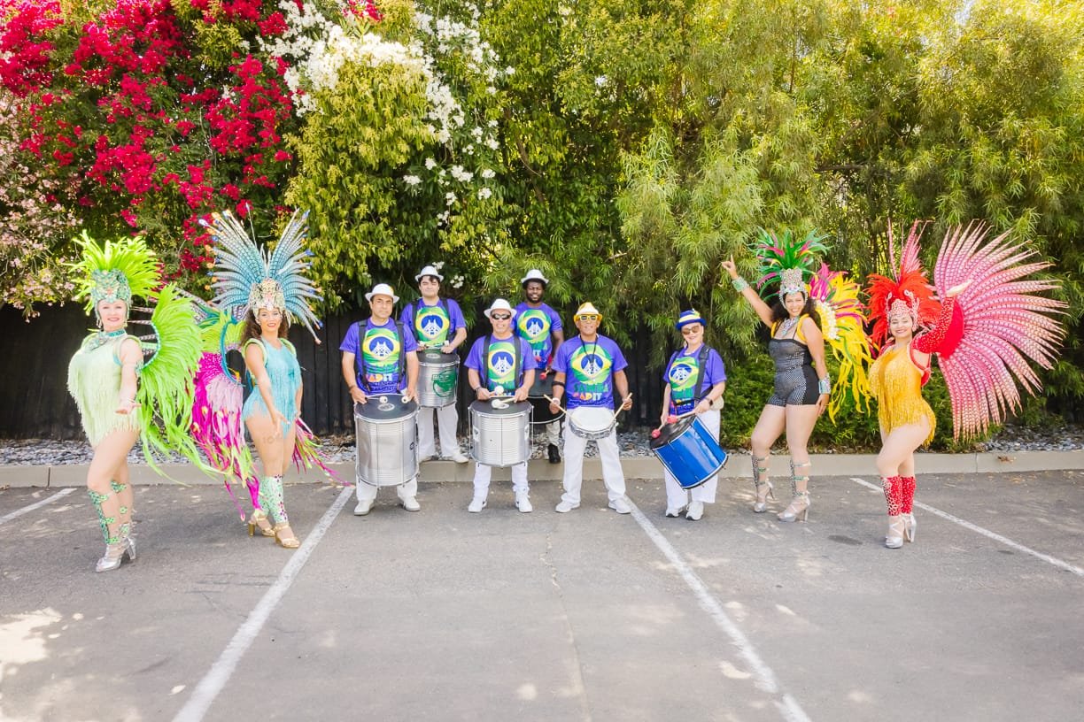 Group of performers in colorful costumes and feathered headdresses posing in a parking lot with trees in the background.  G.R.E.S. unidos da capital of sacramento brazilian samba performing arts group