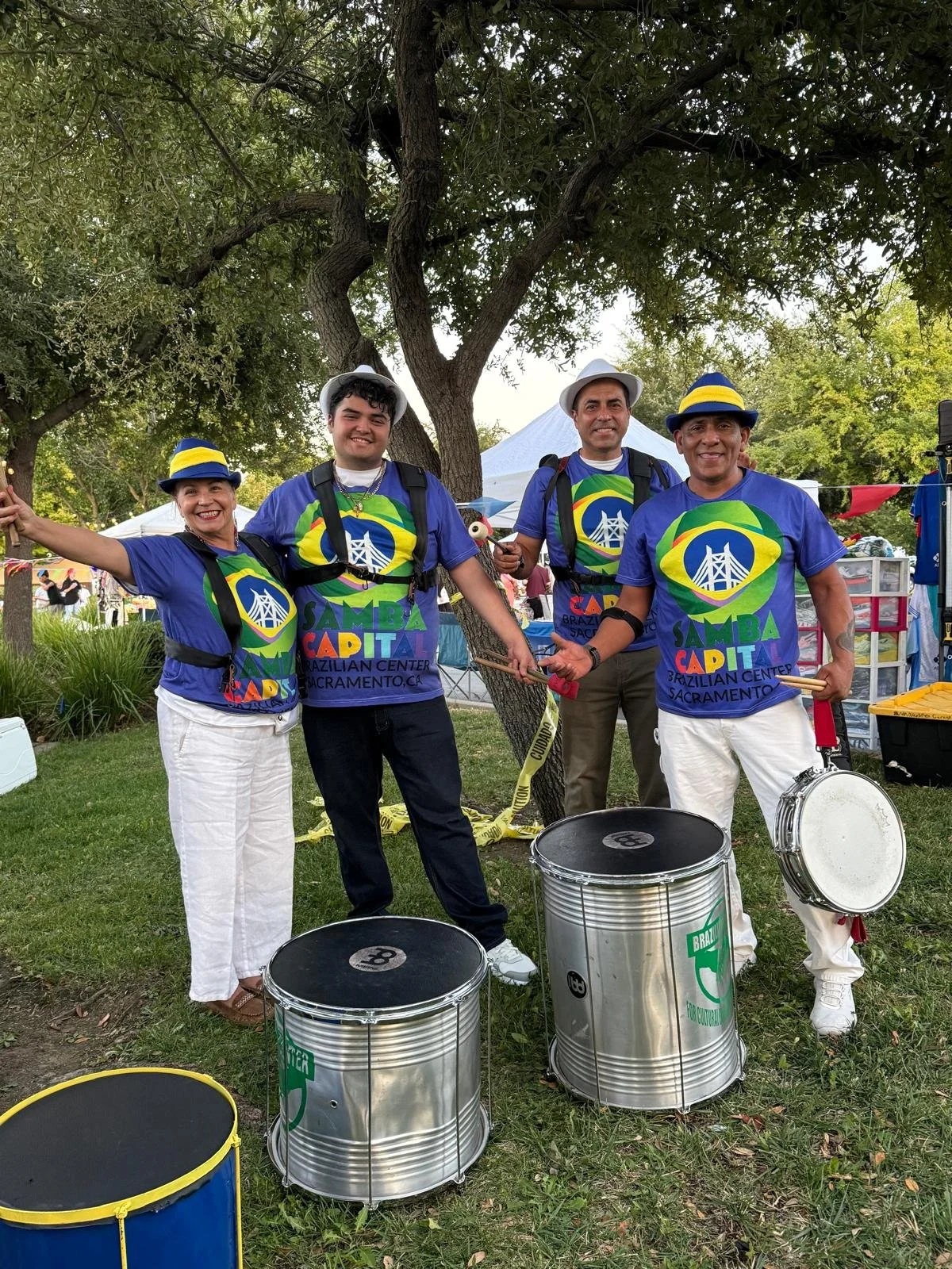 Four people smiling and holding drumsticks, wearing blue shirts with a colorful logo and the words 'Samba Capital' and 'Brazilian Center Sacramento CA.' They are standing on grass in a park under a large tree, with drums in front of them and tents in