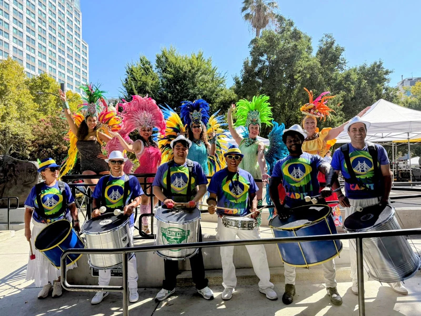 A group of performers dressed in colorful costumes with large feathered headdresses and samba dancers playing drums in front of a park with trees and a tall building in the background.  G.R.E.S. unidos da capital of sacramento brazilian samba 
