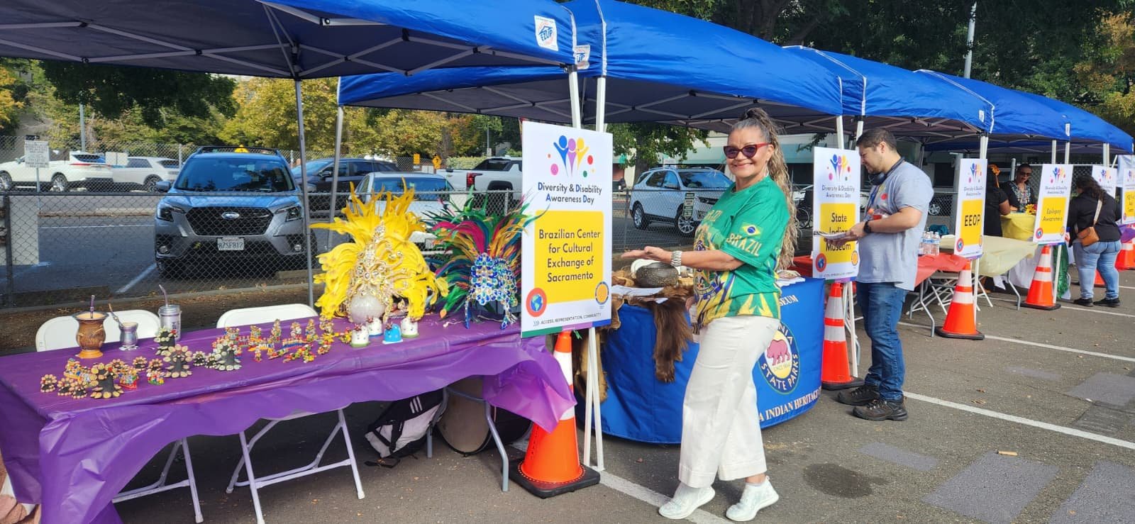 A woman in a green shirt with yellow and blue accents stands next to a table with colorful carnival masks and decorations, part of a booth under a blue tent at a community event celebrating Diversity & Disability Awareness Day.