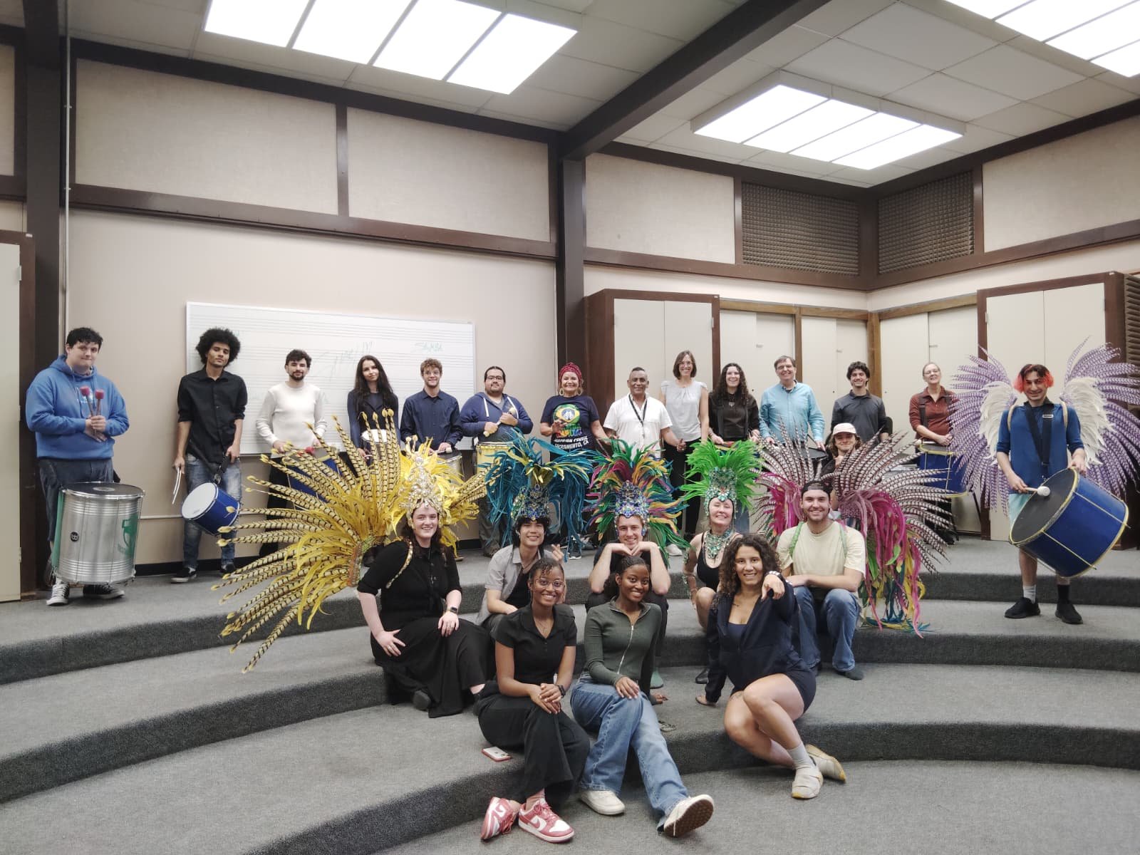 Group of people dressed in costumes with feathered headdresses and drums, posing on a stage in a classroom or auditorium. Samba class in sacramento