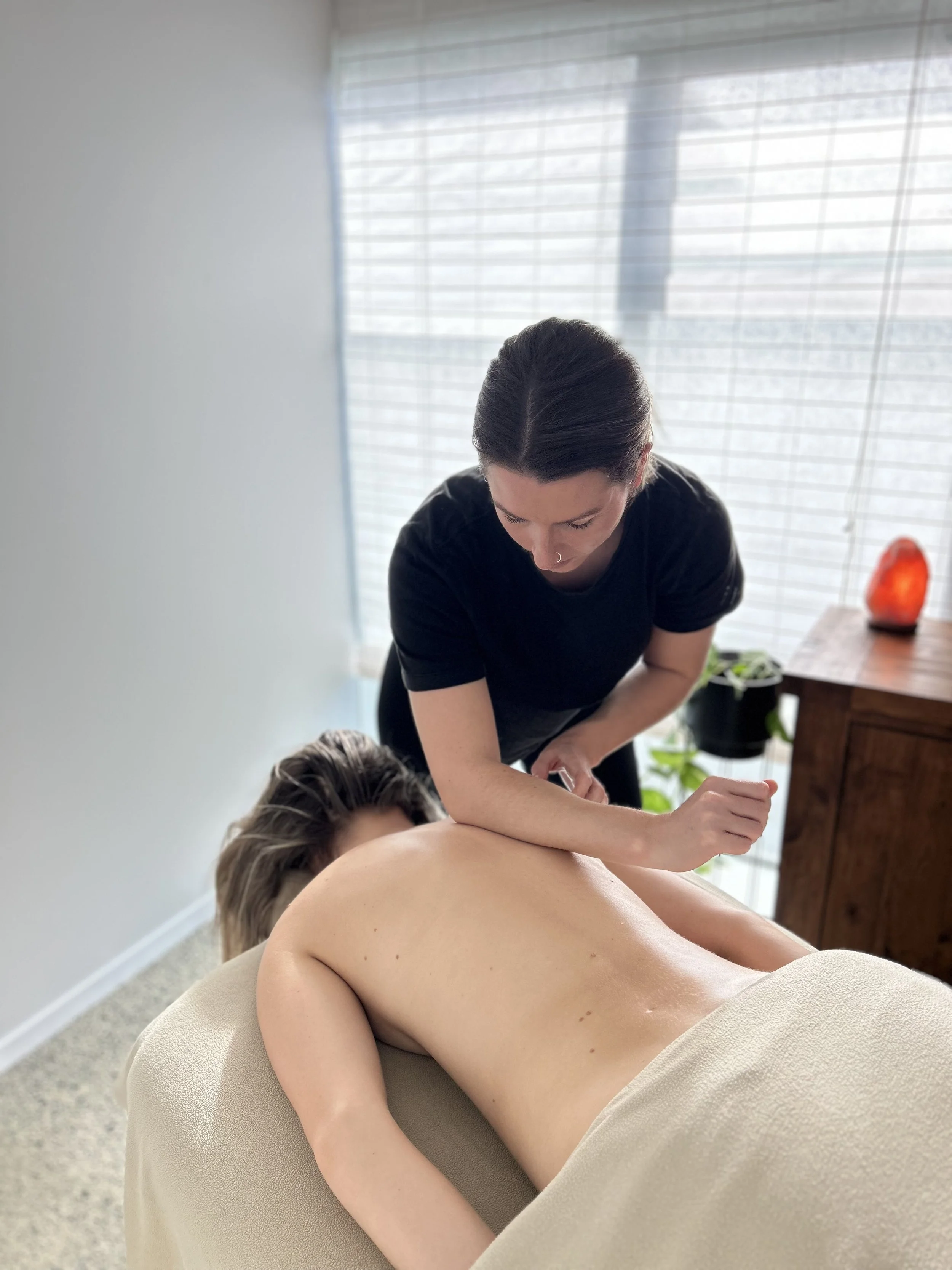 A woman receiving a massage from a massage therapist in a bright room with a window and plants.
