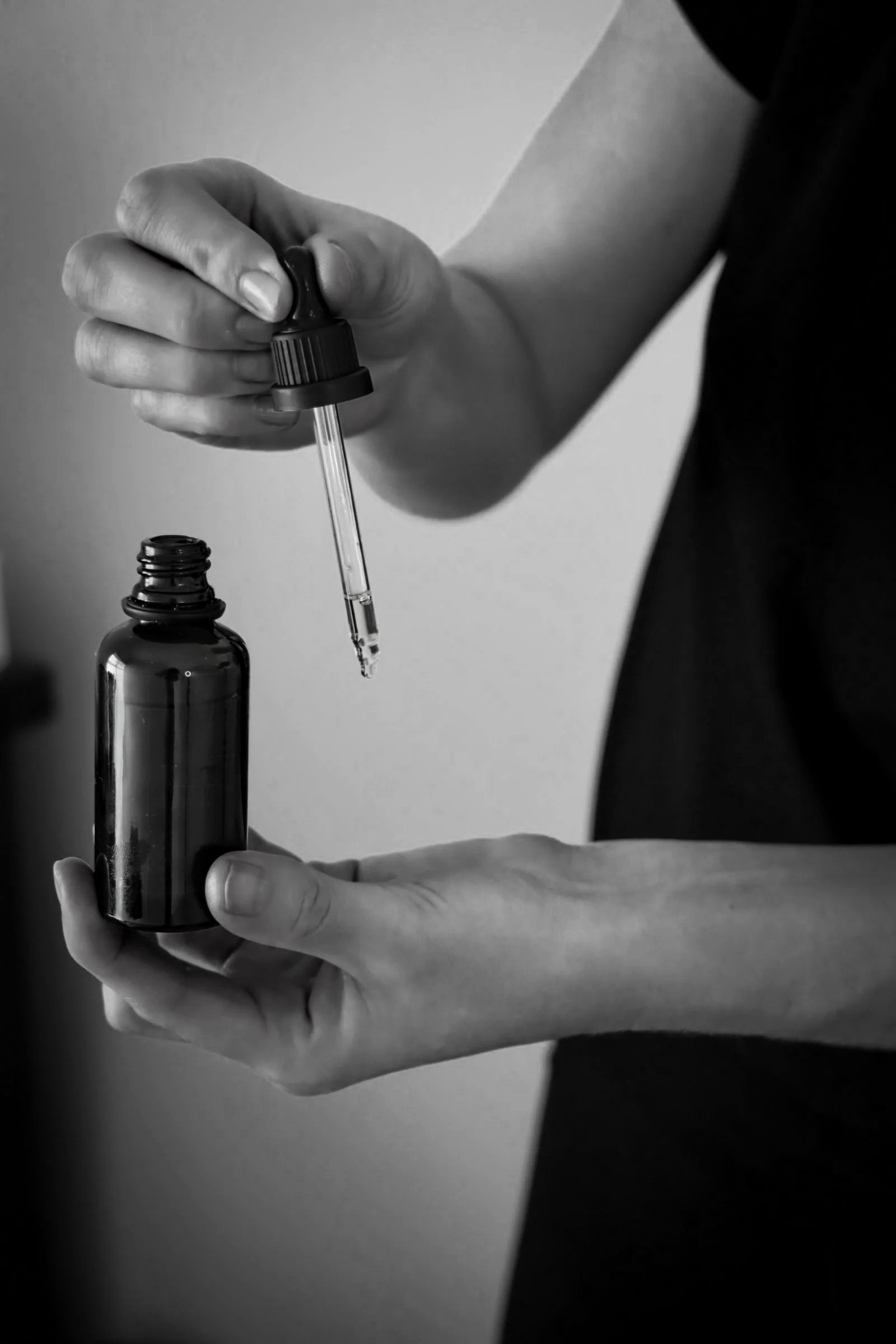 A person holding a small dark glass bottle and using a dropper to transfer liquid into it.
