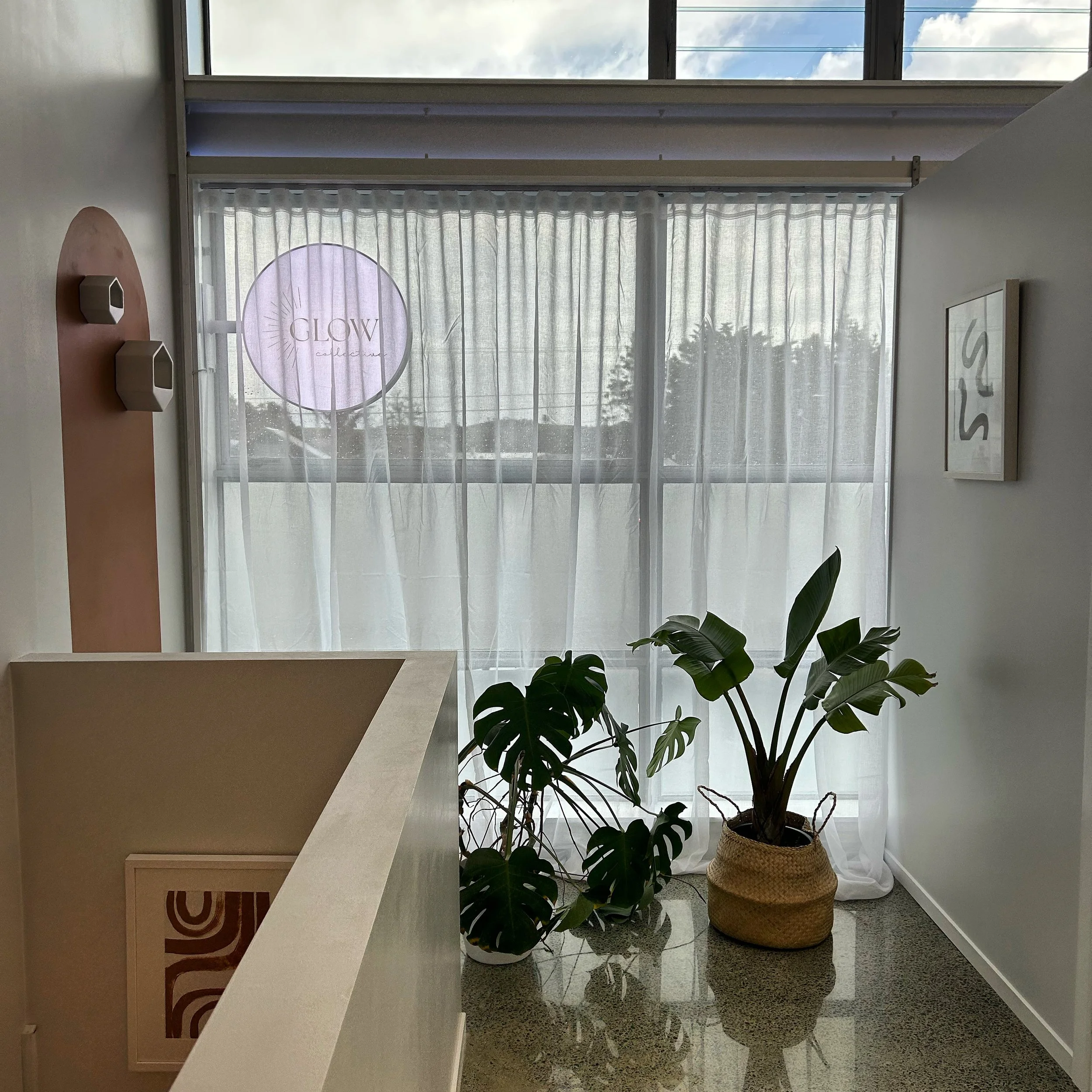 Interior of a cafe with a large window covered by sheer curtains, two potted plants on the floor, a wall with hanging decor, and a sign reading 'GLOW' in the window.