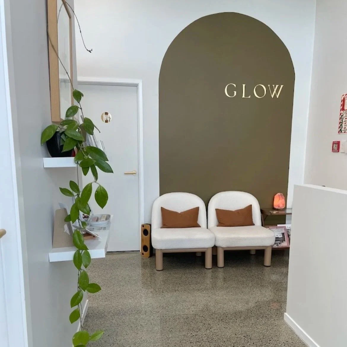 Waiting area with two white chairs with brown pillows, a small table with a salt lamp, a large green accent wall with the word GLOW, a door with a 'No' sign, and a plant on a shelf to the left.