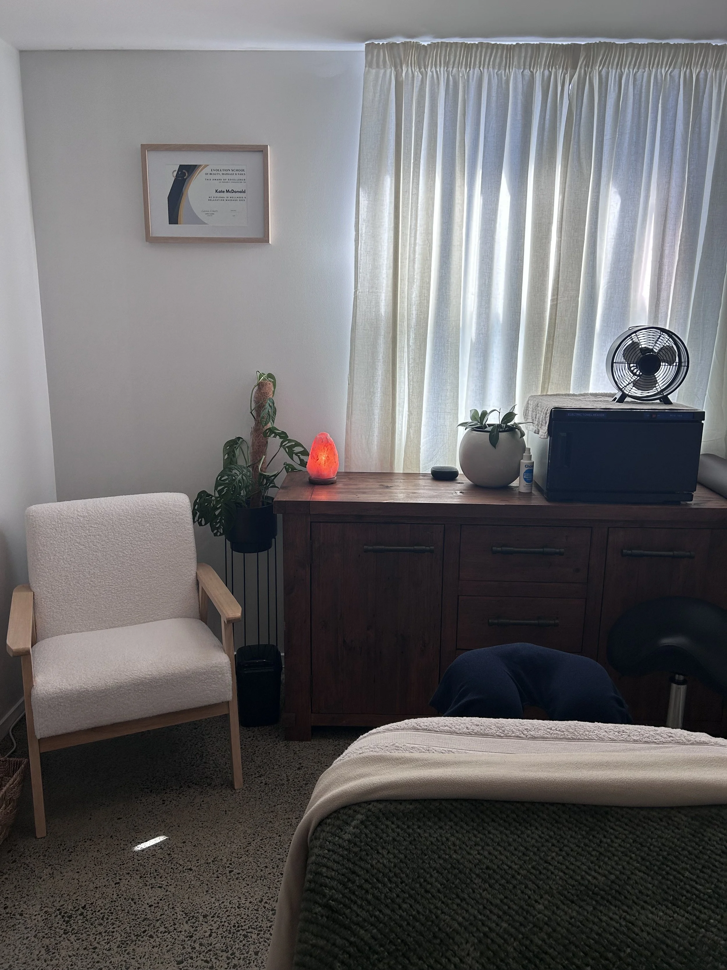 A cozy corner of a room with a white armchair, a wooden dresser, a salt lamp, a potted plant, a framed certificate, a small fan, and a window with light curtains.