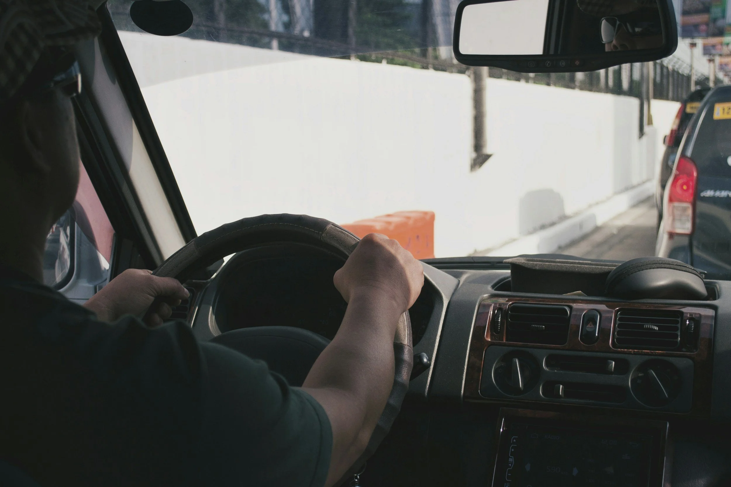 Driver holding steering wheel inside a vehicle on a city street with other cars visible.