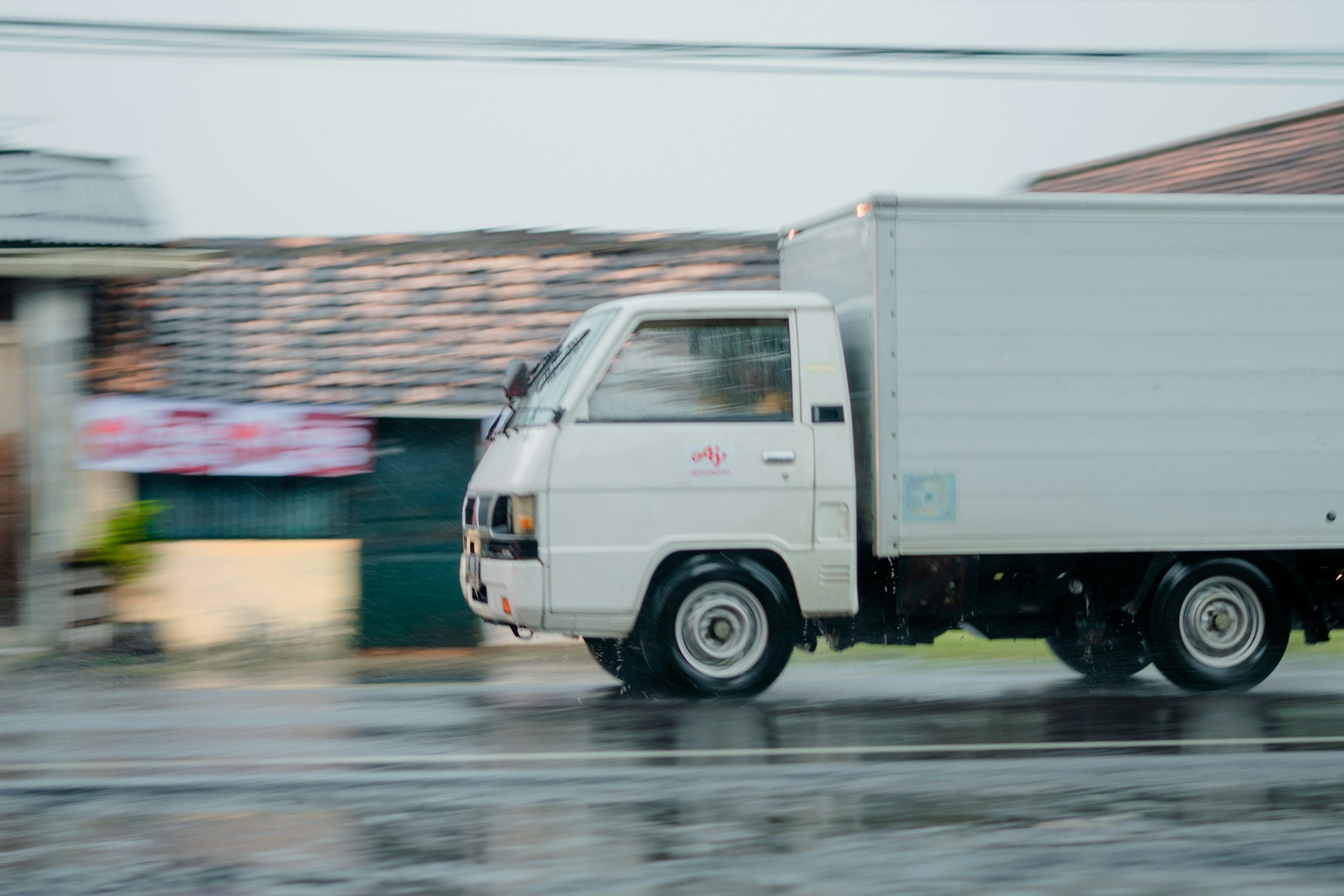 White delivery truck driving on wet road with rain, blurred background of buildings and fence.
