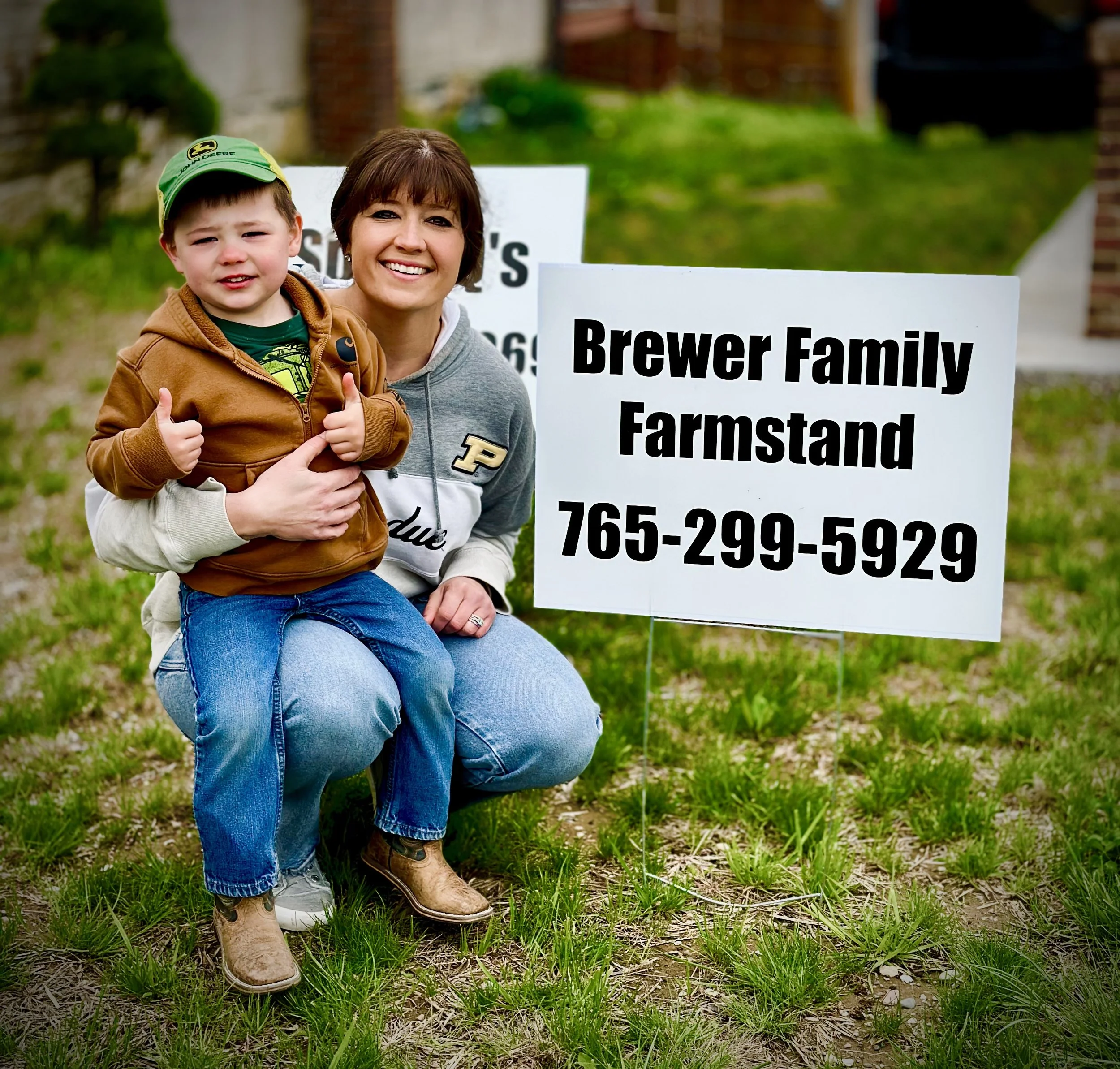Linley Brewer holding her young son beside the Brewer Family Farmstand sign, representing community connection.
