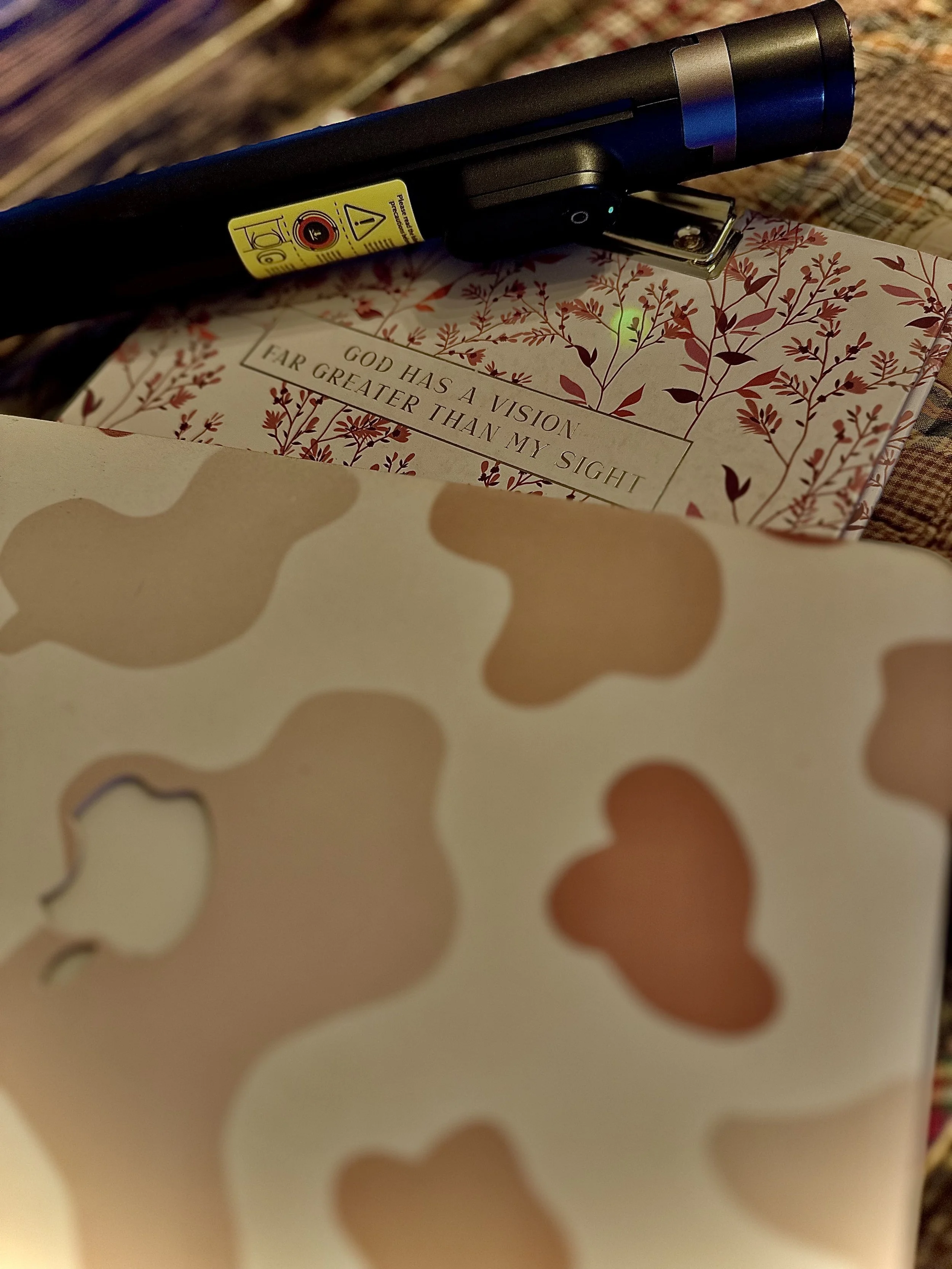 Flat lay of a journal with the phrase 'God has a vision far greater than my sight,' alongside a handheld camera tripod and patterned laptop.