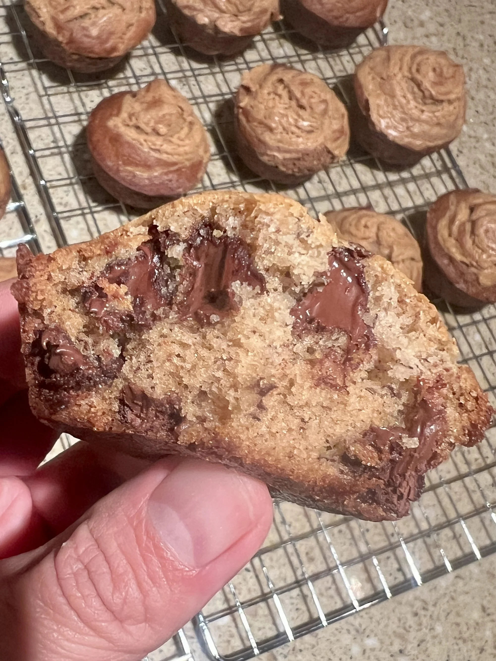 Close-up of a chocolate chip banana muffin cut in half, showing the melted chocolate pockets and soft, moist crumb, with more muffins cooling on a wire rack in the background.