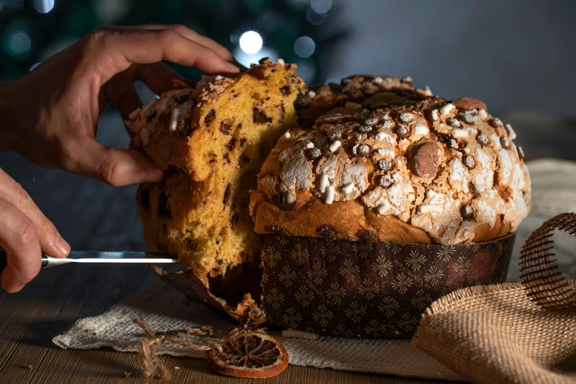 Hands slicing into a tall Italian panettone loaf topped with sugar and almonds, revealing its soft, golden interior with chocolate pieces.