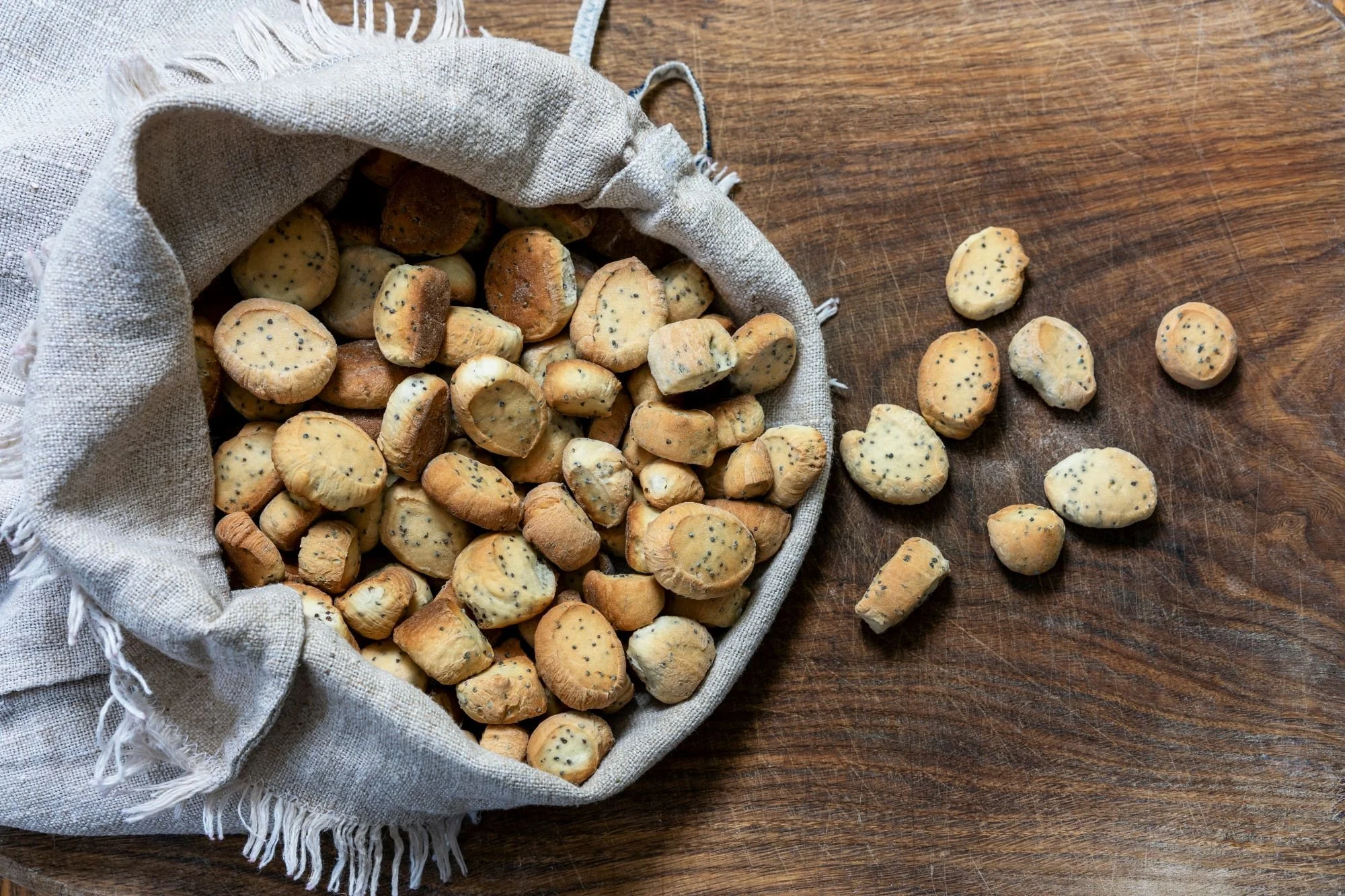A linen sack filled with Lithuanian kūčiukai, small poppy seed Christmas Eve bread bites, with several pieces scattered on a wooden table.