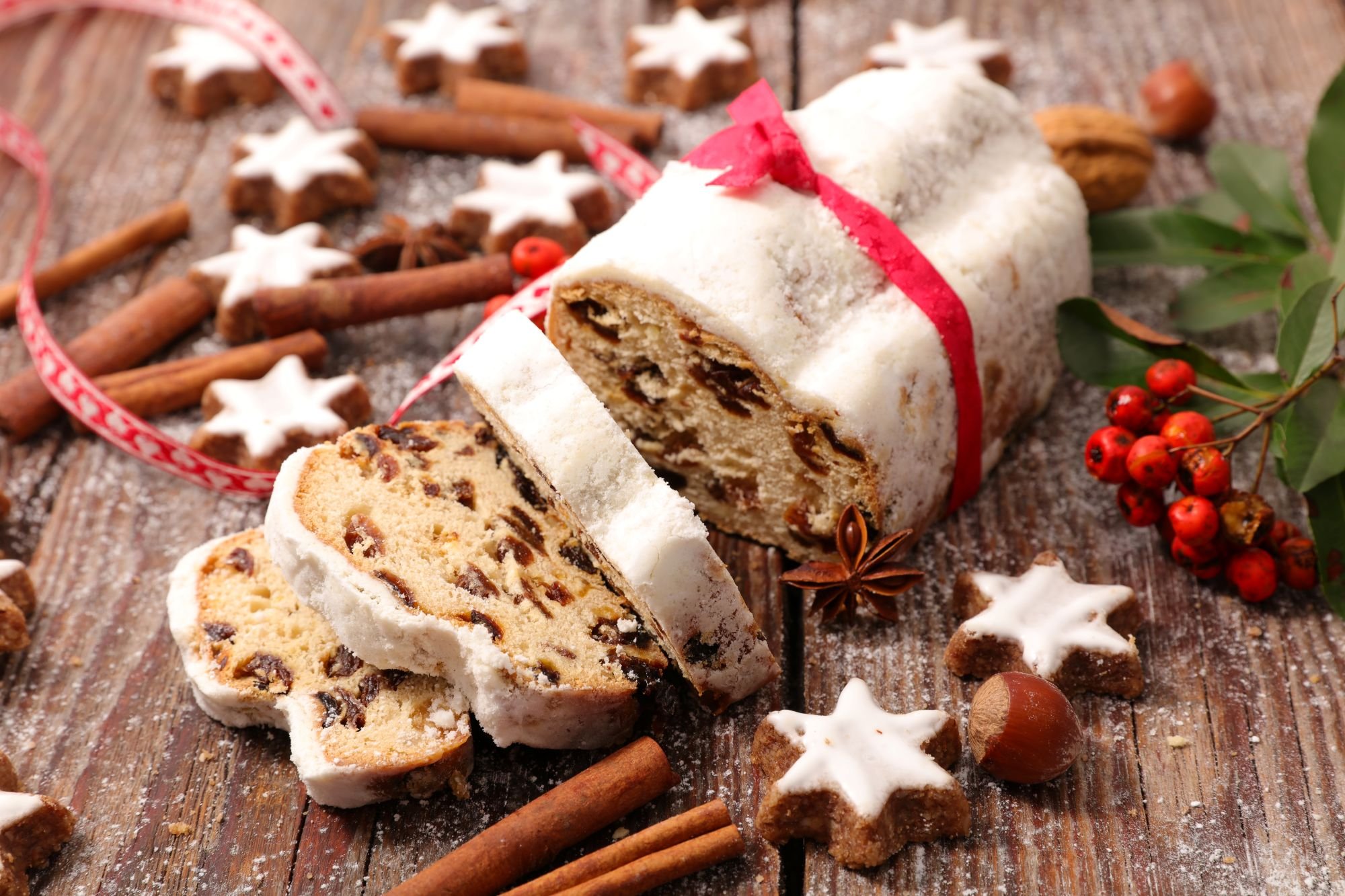 A sliced German stollen loaf wrapped with a red ribbon, dusted in powdered sugar and filled with dried fruit, surrounded by cinnamon sticks, star-shaped cookies, nuts, and festive berries on a rustic wooden table.