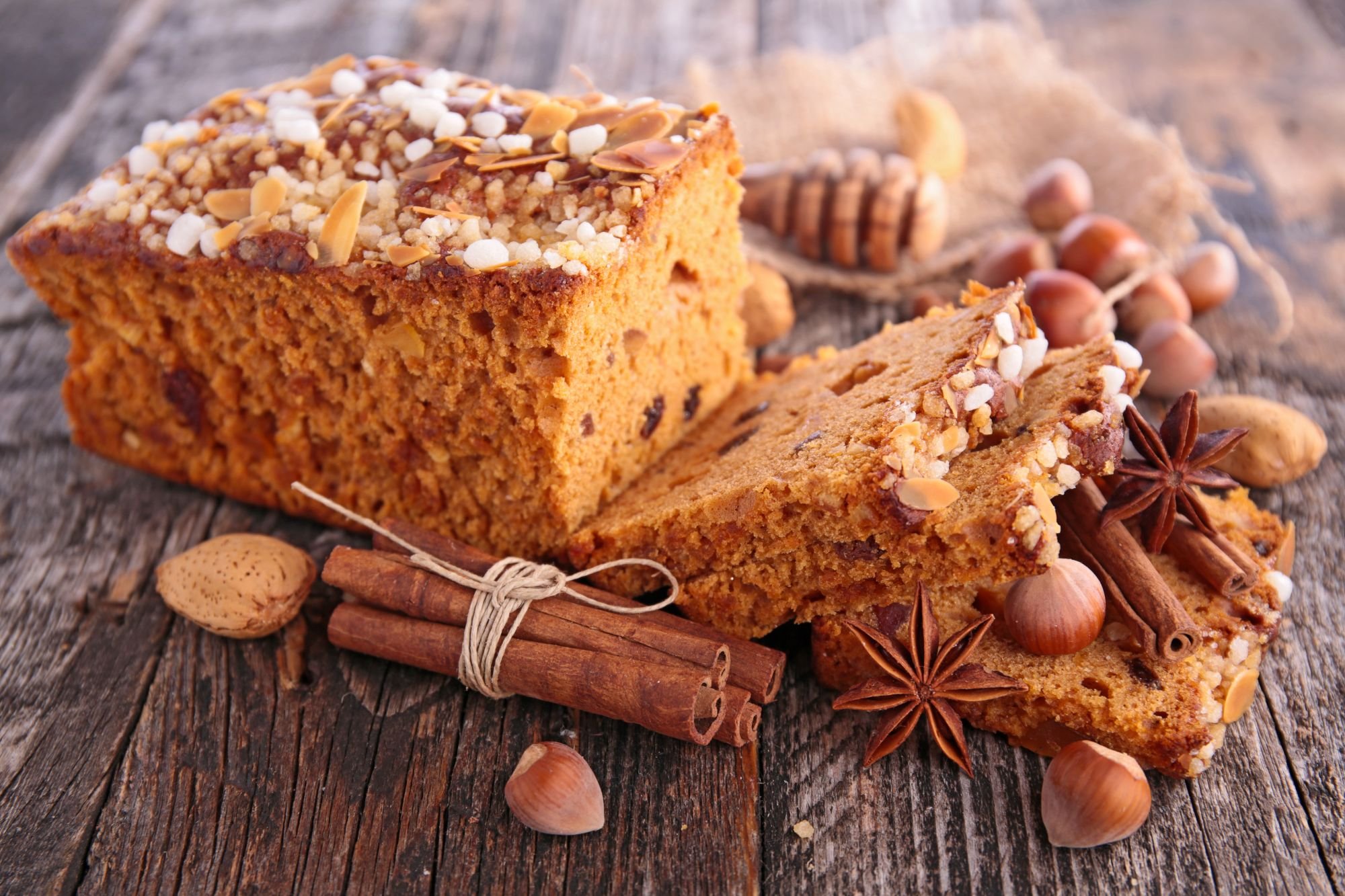 A sliced loaf of gingerbread topped with pearl sugar and almonds, surrounded by cinnamon sticks, star anise, and hazelnuts on a rustic wooden surface.