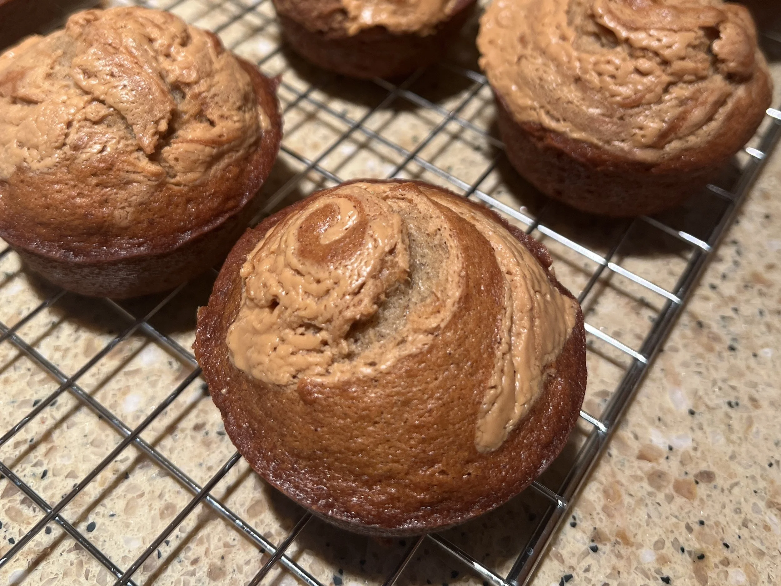Banana muffins with a peanut butter swirl on top cooling on a wire rack, showing the golden brown tops and smooth, creamy swirl pattern.