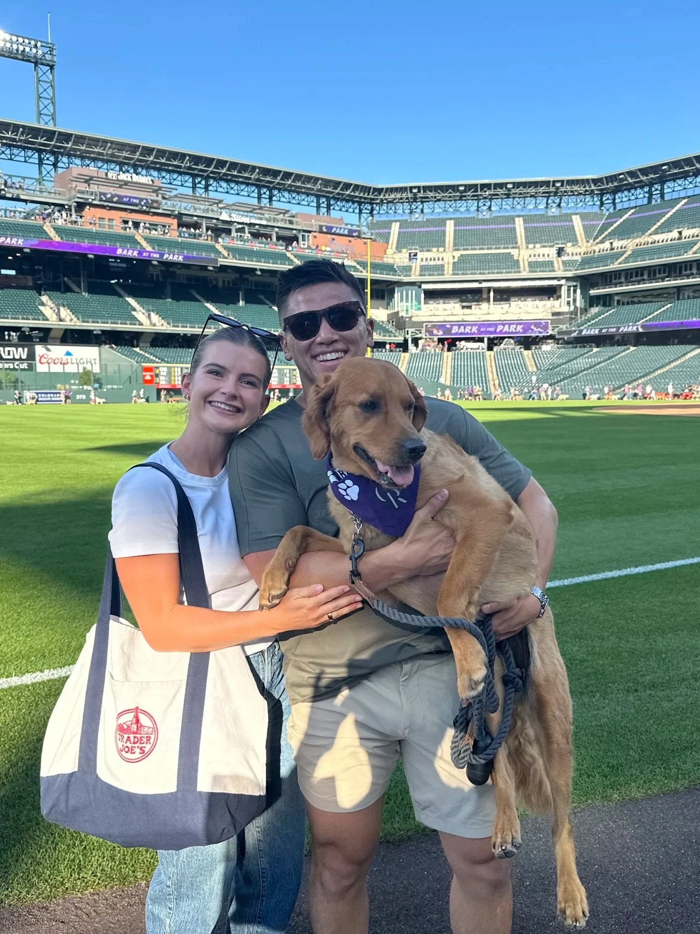 A happy couple, a woman and a man, are standing on a baseball field with their dog, a golden retriever, at a stadium. The man is holding the dog, and the woman is smiling with her arm around him. Chicago Dental Anesthesia sedation Illinois