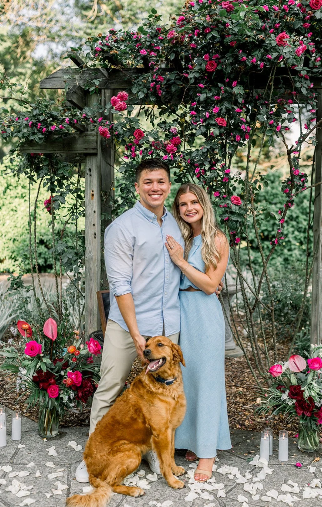 A happy couple with a dog standing in front of a floral arch with pink roses, surrounded by vases of flowers, candles, and petals on the ground, outdoors. Chicago Dental Anesthesia sedation Illinois