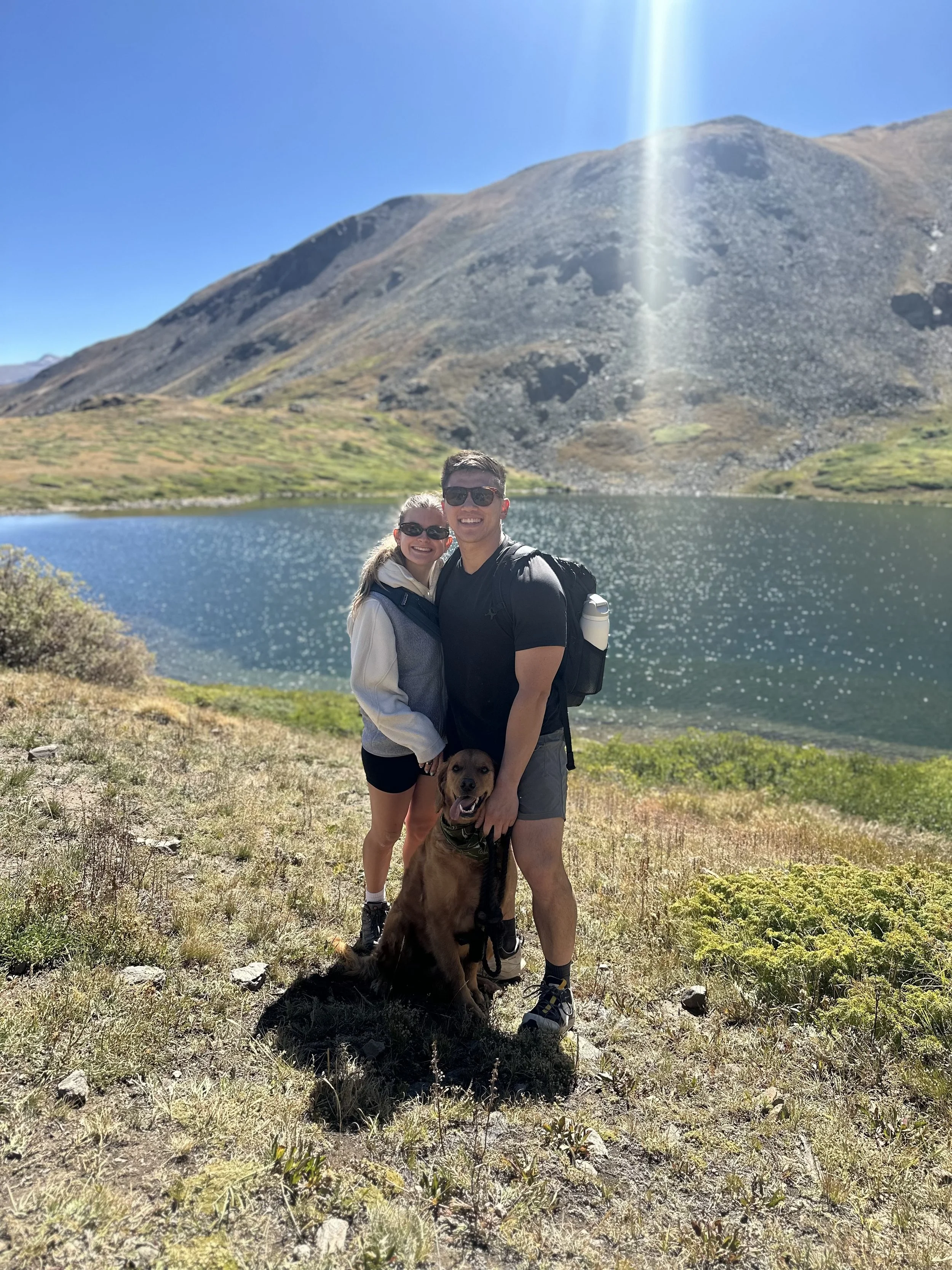 A happy couple with a dog standing in front of a lake with mountains in the background on a sunny day. Chicago Dental Anesthesia sedation Illinois