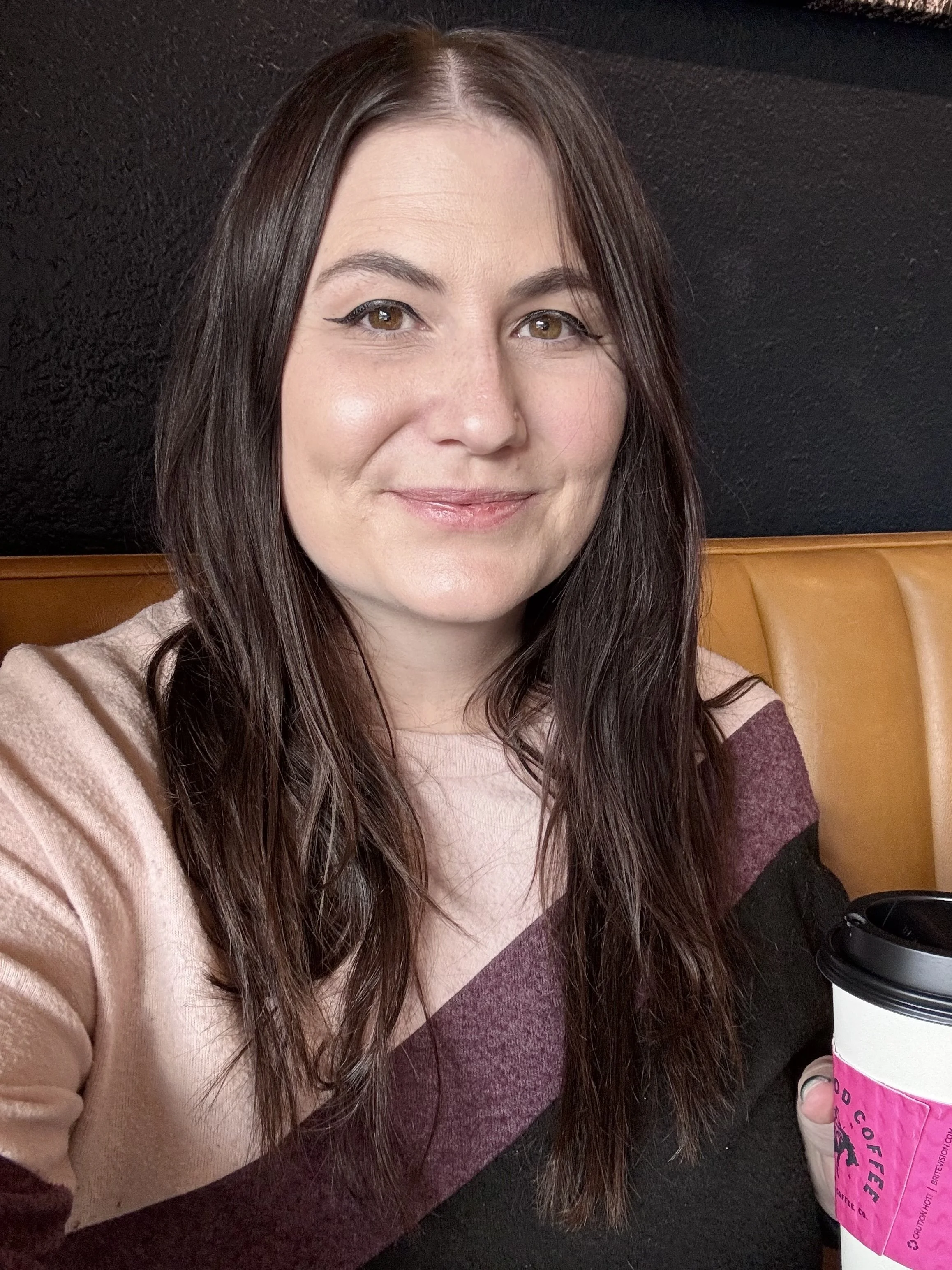 A woman with brown hair and light makeup sitting in a booth at a coffee shop, holding a cup of coffee.