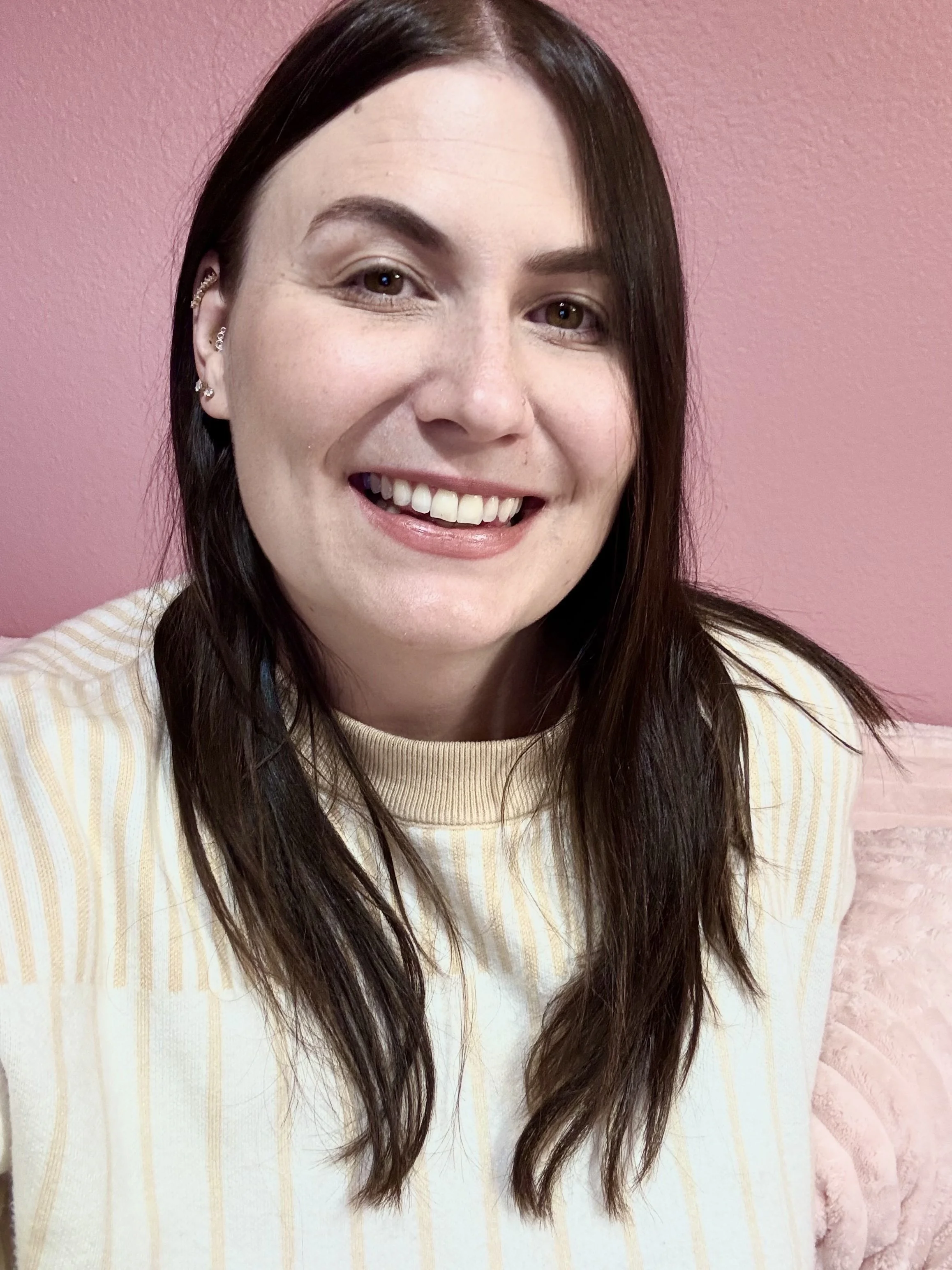 A young woman with long dark brown hair and brown eyes smiling at the camera, wearing a cream and yellow striped sweater, in front of a pink wall.