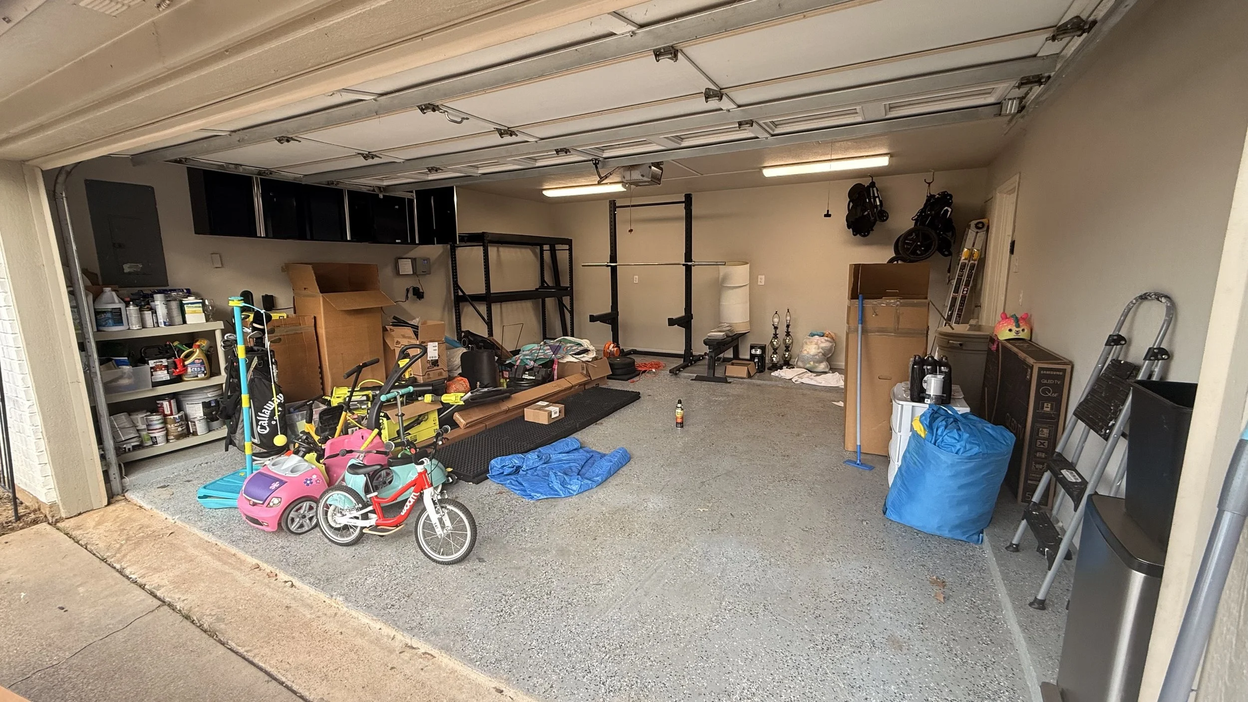 Open garage with various items including children's toys, workout equipment, shelving, storage boxes, ladders, and bottles, with some items on the ground and shelves.
