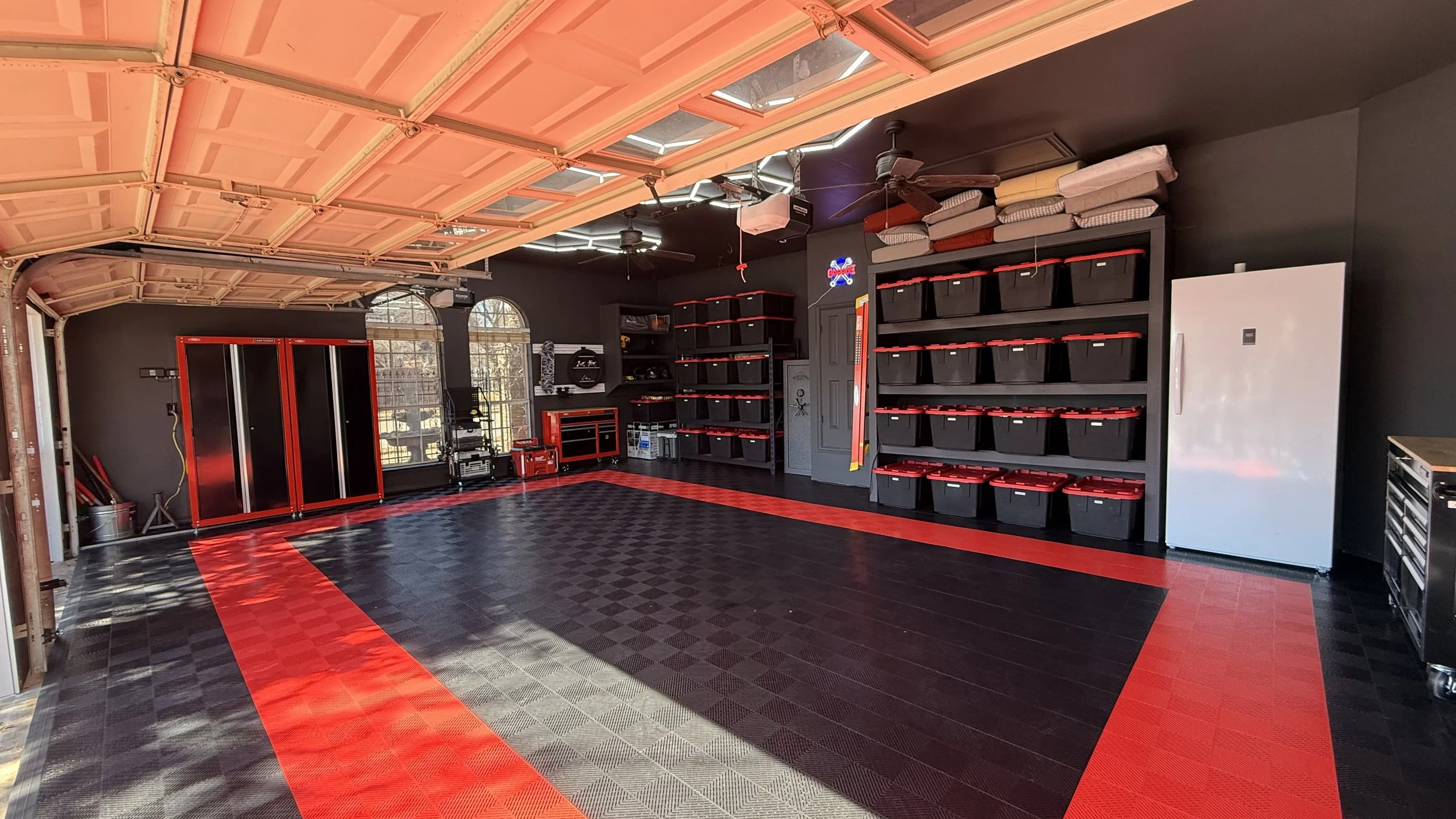 Spacious garage with black walls and red accents, organized with black storage bins on shelves, a white refrigerator, and a black and red tool chest. The garage door is open, allowing sunlight inside.