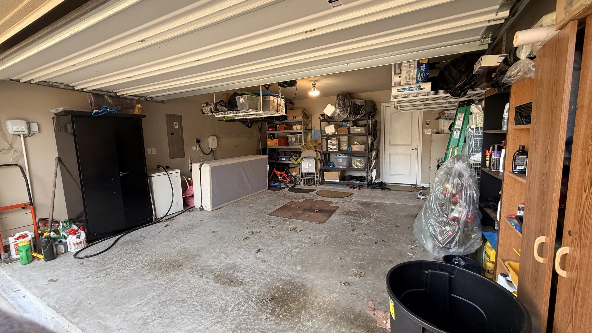 Inside a garage with an open door, showing various tools and storage shelves, a bicycle, a ladder, and a large black trash bin.