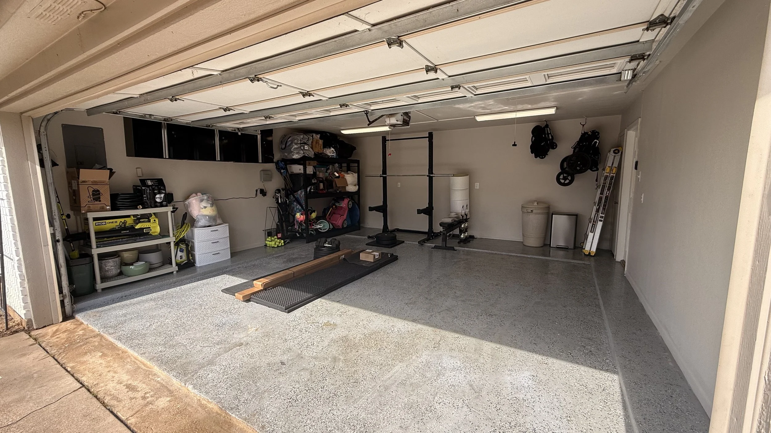A spacious garage with various storage shelves, workout equipment, and household items organized along the walls. The garage door is open, revealing a sunny day outside.