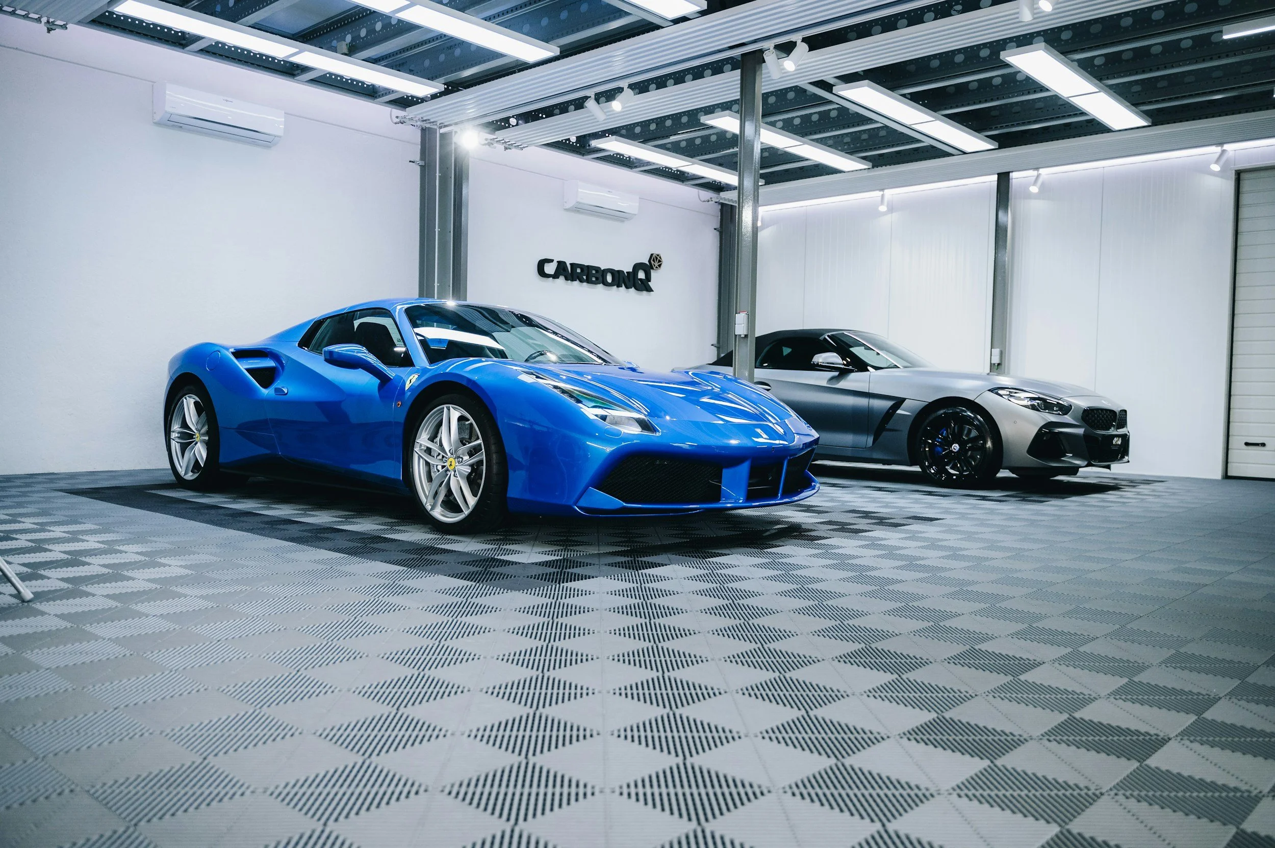 Two luxury sports cars, a blue Ferrari and a silver BMW i8, parked side by side inside a modern showroom with a sign that says 'CARBONQ' on the wall.