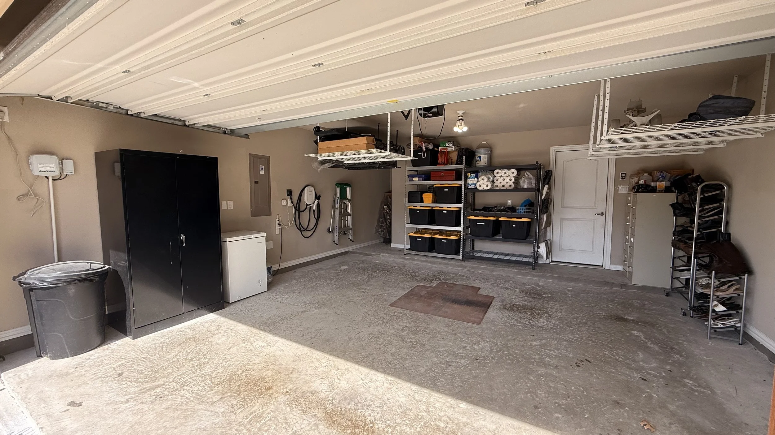 Empty garage with shelves containing bins and storage containers, ladder, wall-mounted hose reel, and a trash can.