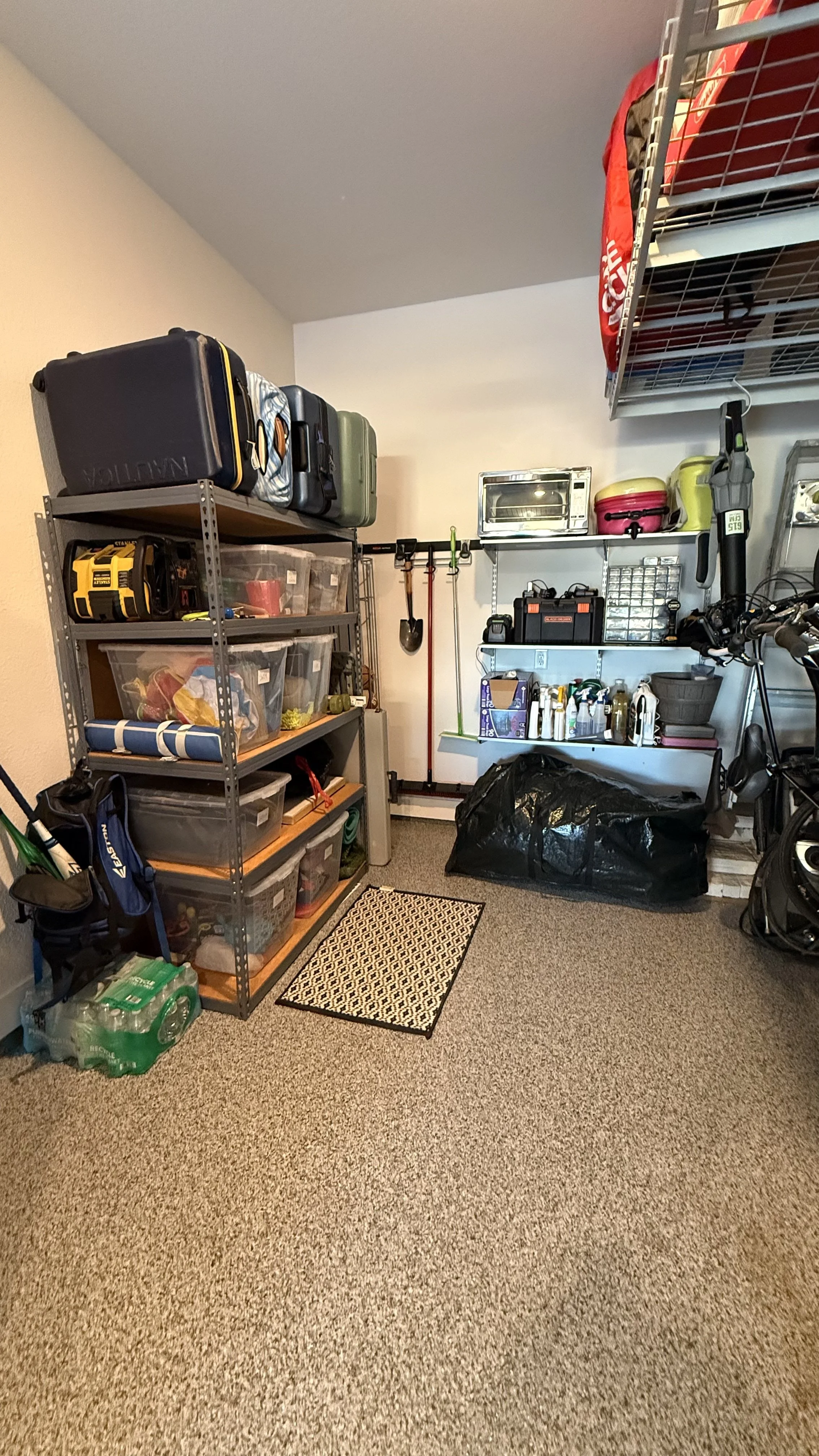A garage storage area with metal shelving units filled with bins, boxes, and tools, a black garbage bag, and a step stool on a carpeted floor.