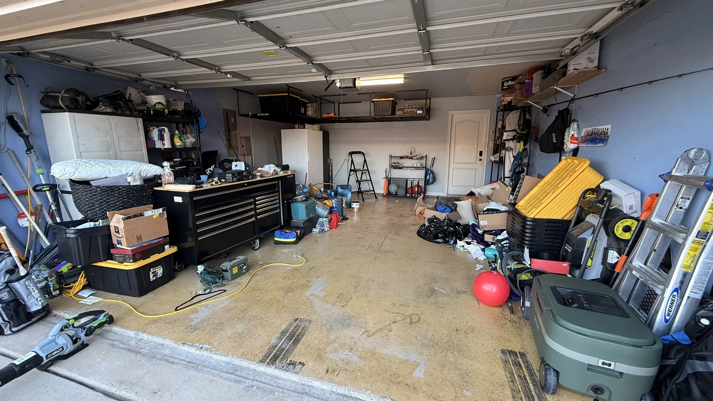 A cluttered garage with various tools, storage containers, and miscellaneous items scattered on the floor and shelves.