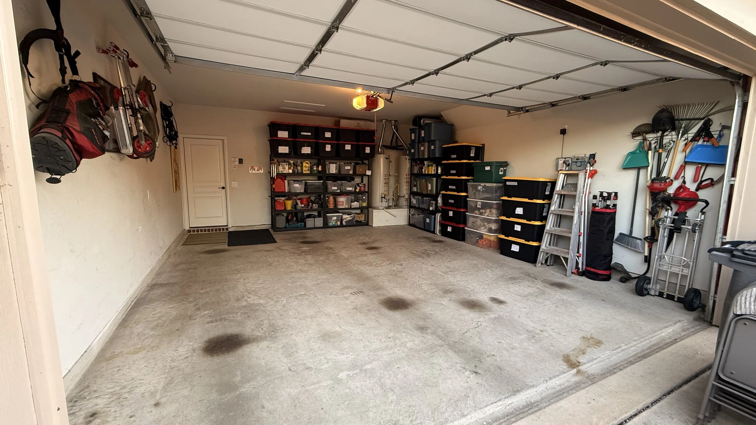 Empty garage with organized shelves filled with storage bins, tools, and gardening supplies; wall-mounted yard tools on the left; a ladder and rolling cart on the right; concrete floor with some stains.