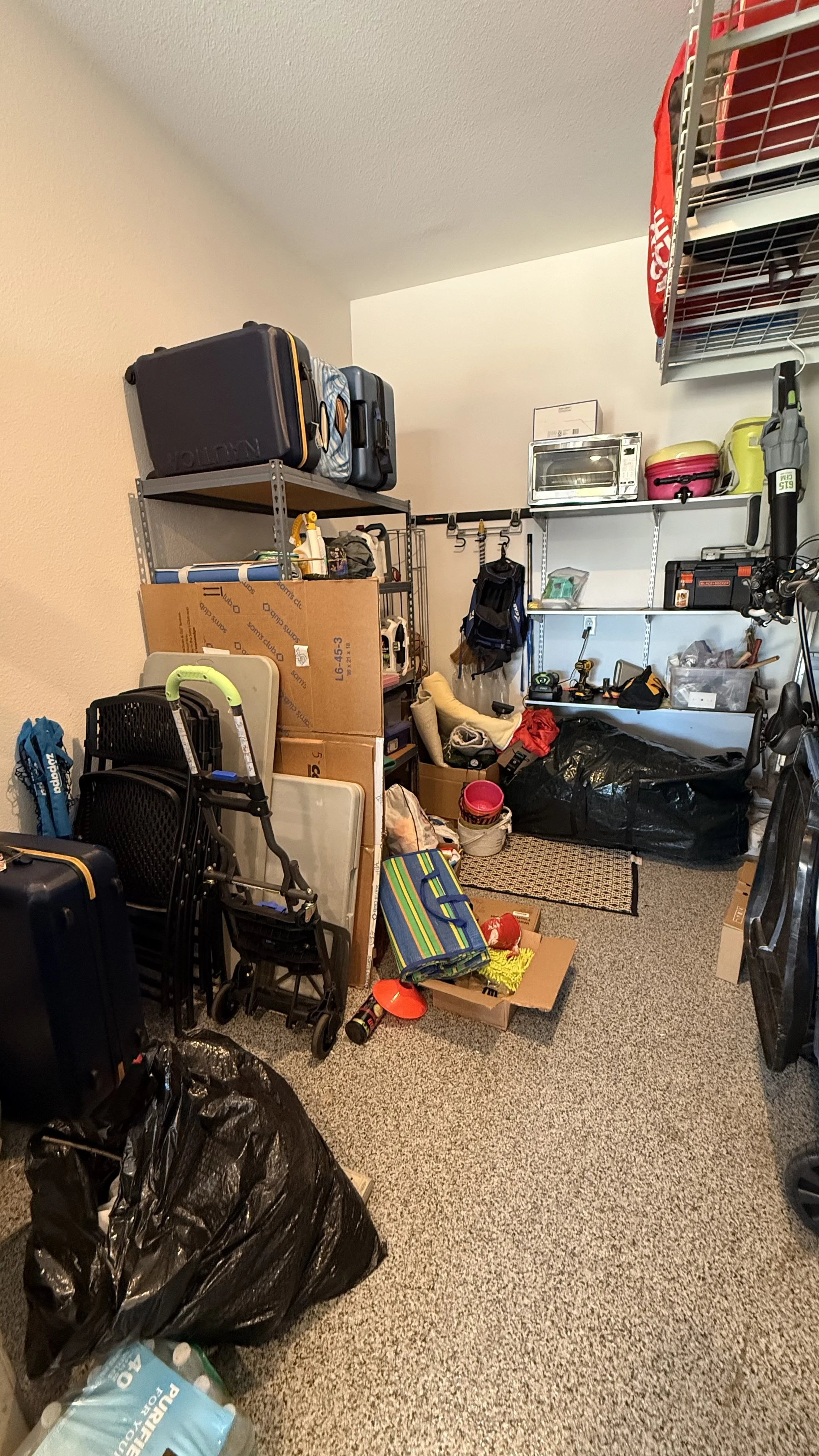A cluttered storage room with shelves filled with boxes, travel luggage, and miscellaneous items, including a folding chair, a small shopping cart, and various bags and household supplies.