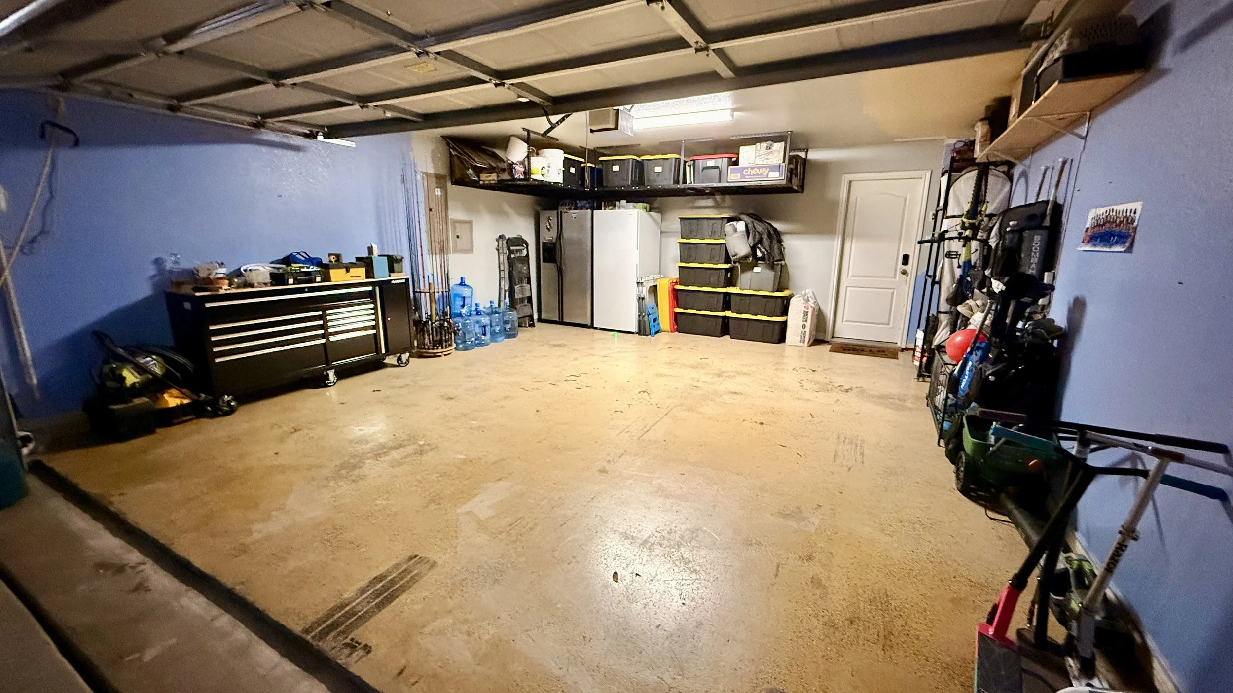 A garage with blue walls and a concrete floor, containing storage shelves, a large refrigerator, water bottles, and various tools and equipment.