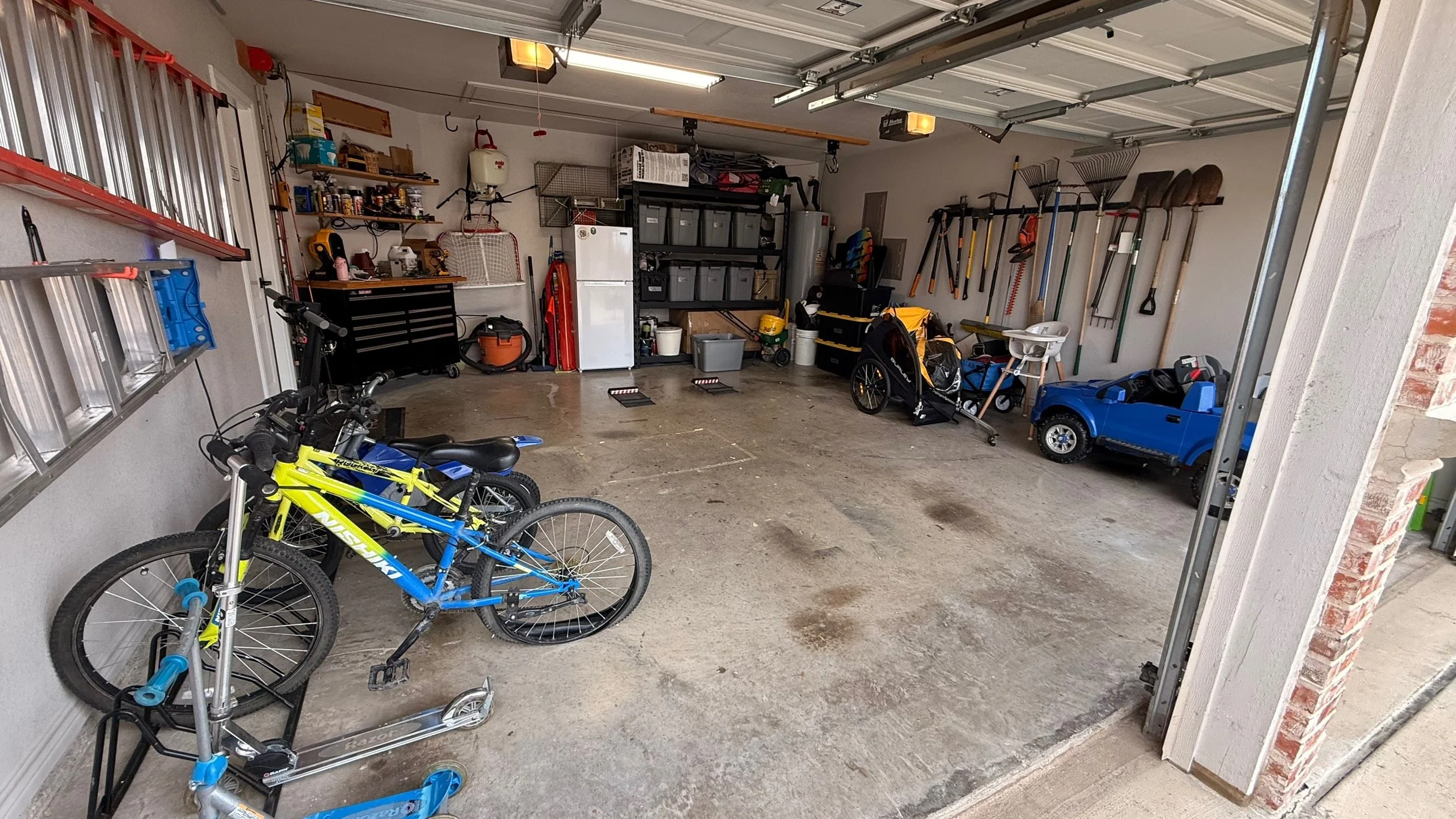 A spacious garage with bicycles, various tools, storage shelves, and a toy car. The garage has a concrete floor and organized storage areas on the walls and shelves.
