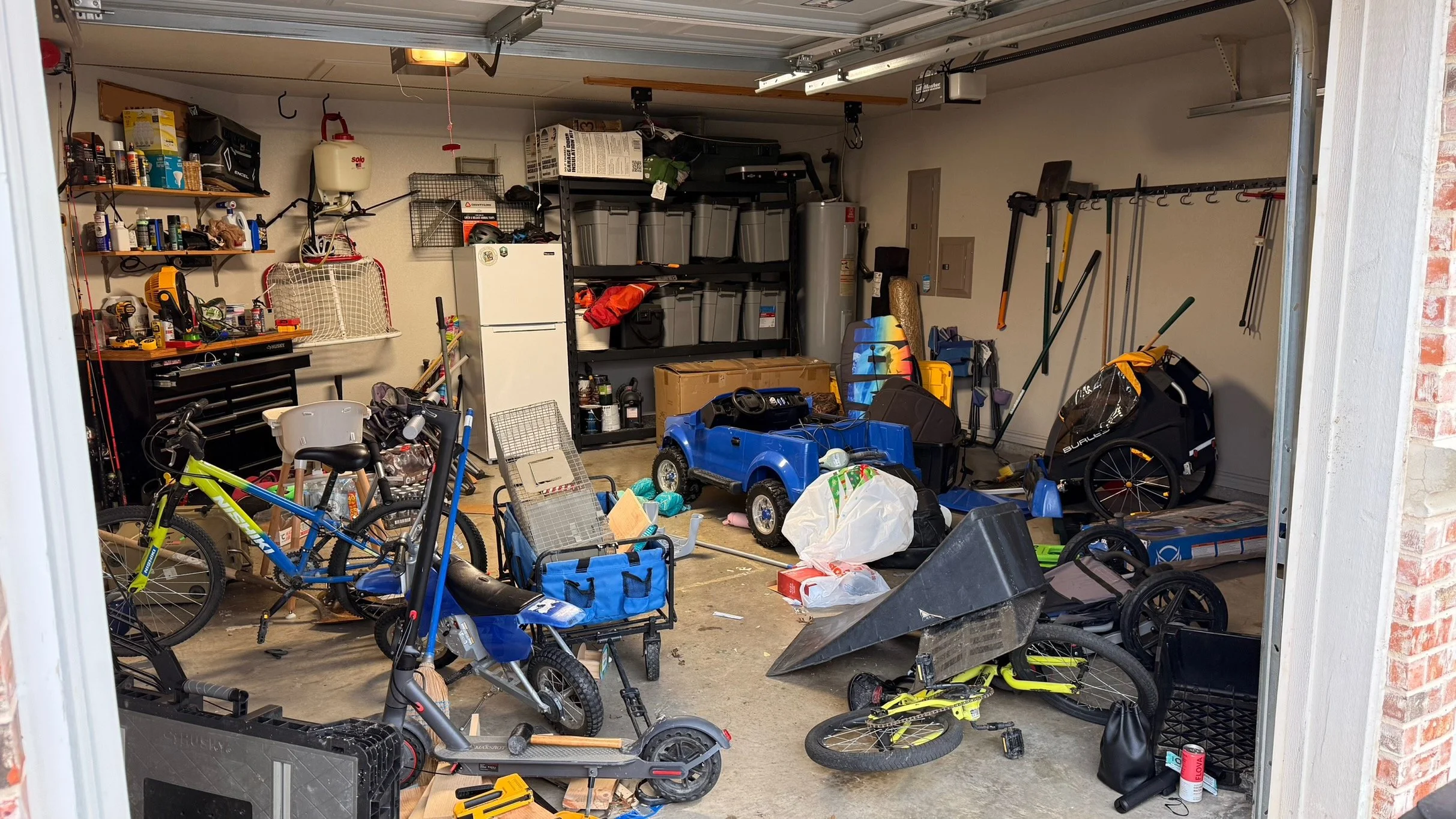 A cluttered garage filled with toys, bicycles, tools, storage bins, a refrigerator, and various gardening tools and equipment.
