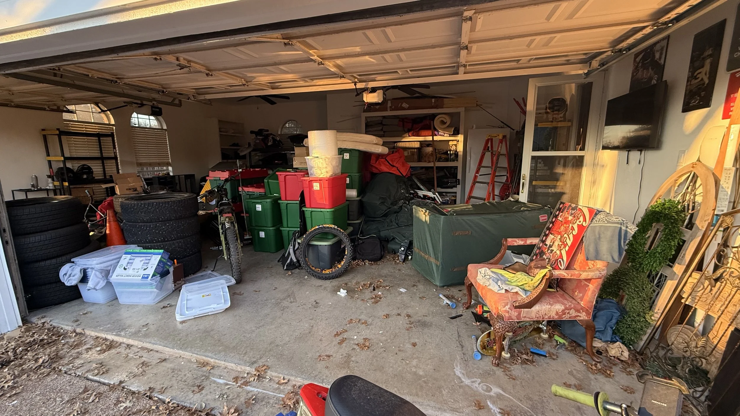 Disorganized garage with stacked tires, plastic storage bins, a bicycle, a chair with a cloth, a TV mounted on the wall, and various tools and supplies.