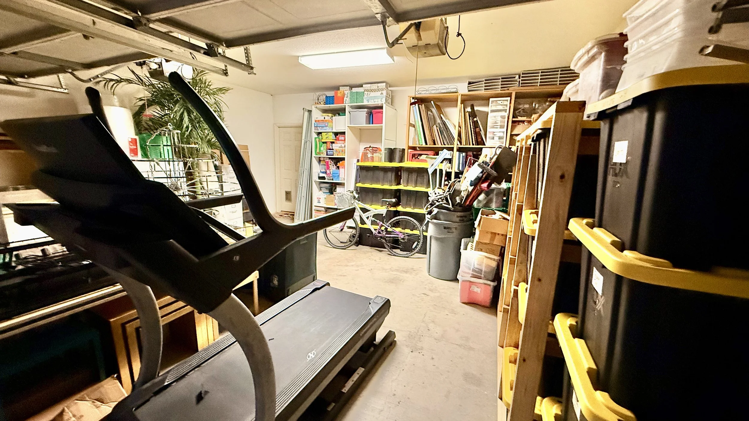 A cluttered garage with a treadmill, stacked black storage bins with yellow lids, bikes, shelves filled with books, boxes, and various tools, and a large potted plant in the background.