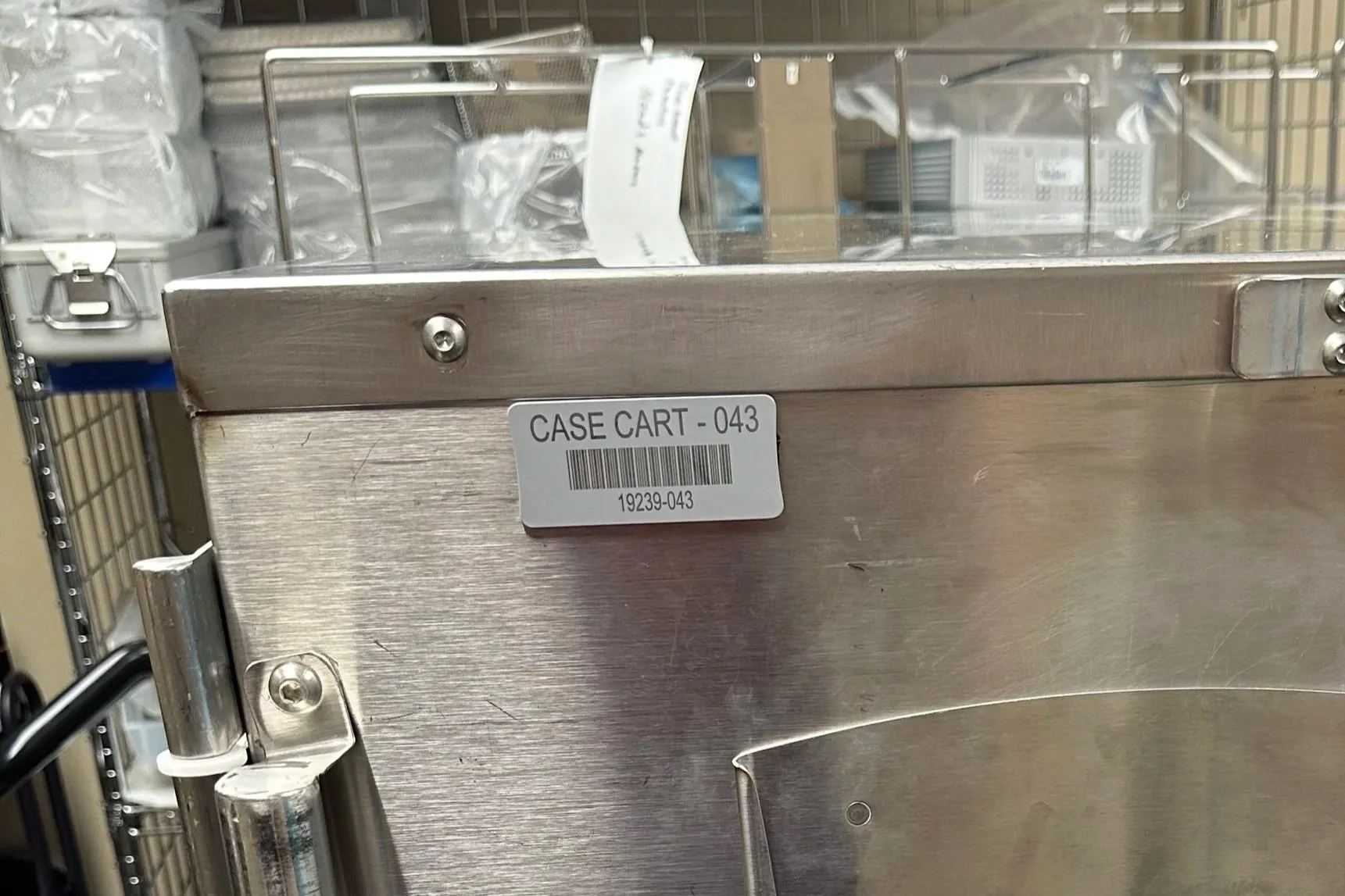 A stainless steel case cart with a label reading 'CASE CART - 043' and a barcode, in a storage or hospital supply room.