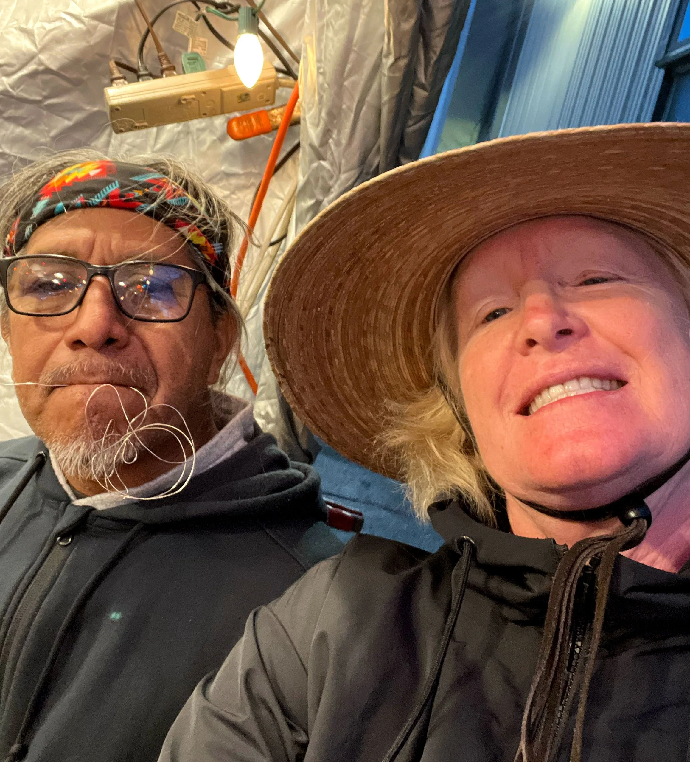 Two people taking a selfie, one wearing glasses and a colorful headband, the other wearing a large straw hat, both in outdoor jackets, with a makeshift outdoor or work area in the background.